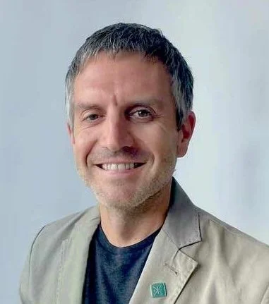 A man with short dark hair smiling, wearing a beige blazer over a dark shirt, against a light background.
