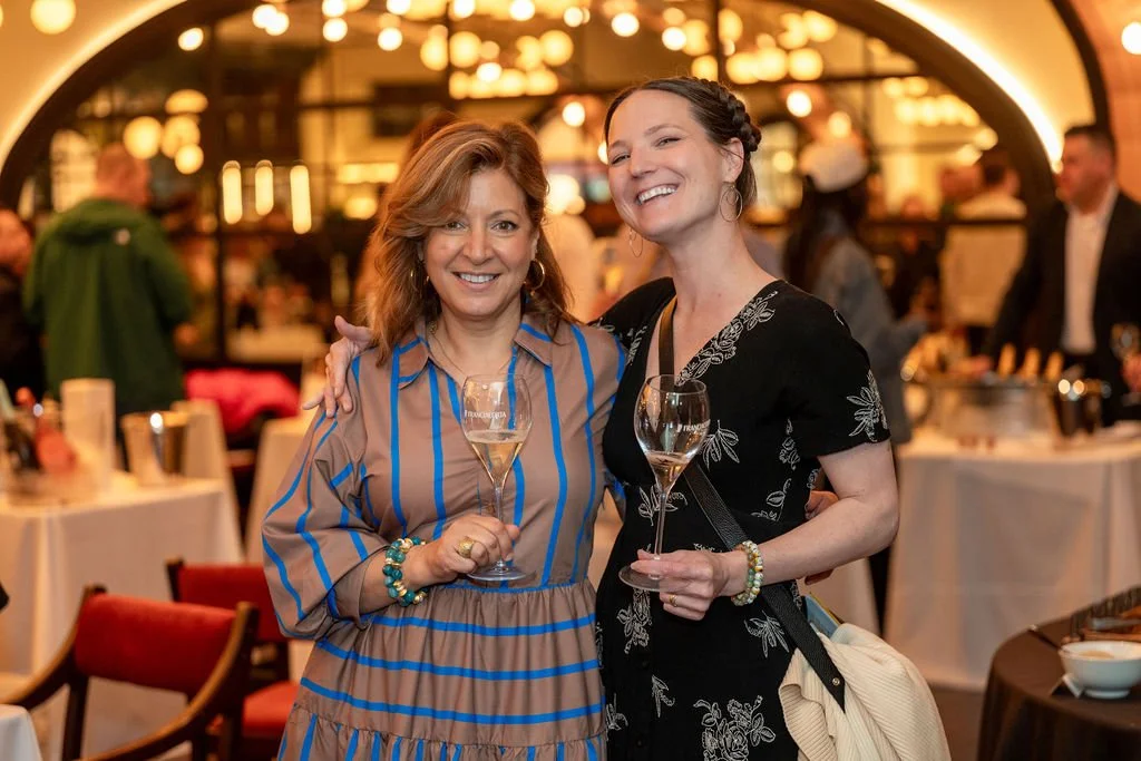 Two women smiling and holding glasses of white wine at a social event in a warmly lit restaurant or bar, with other guests and tables in the background.