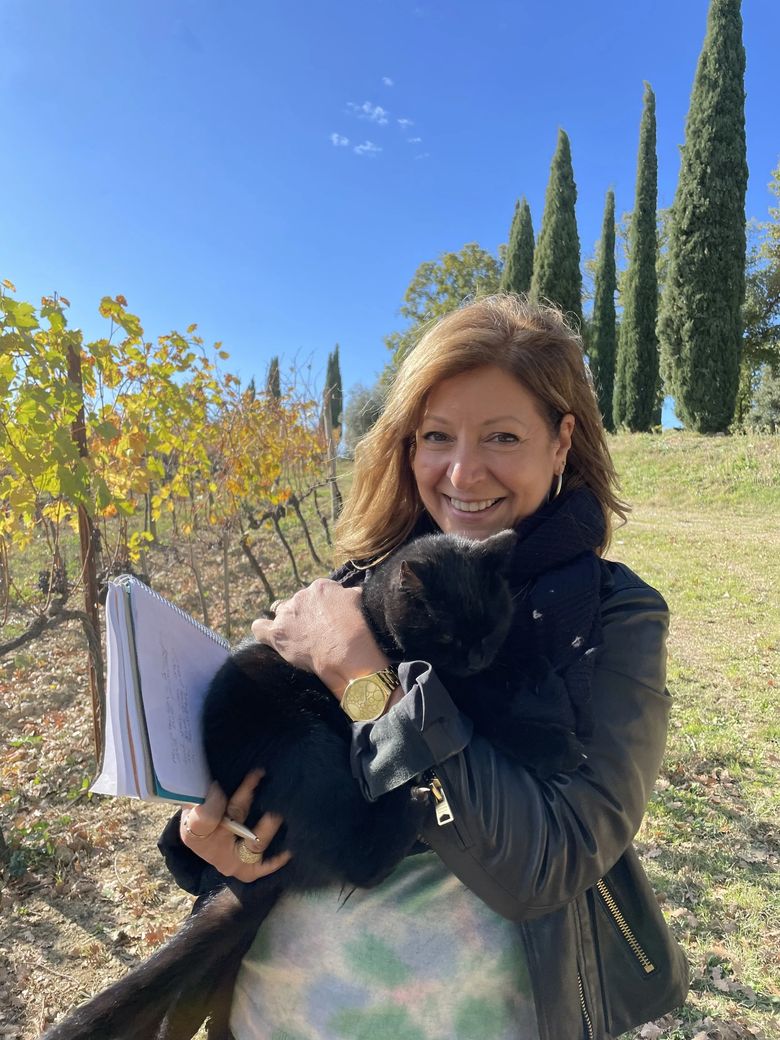 A woman smiling outdoors, holding a black cat and a notebook, with a backdrop of tall cypress trees and a clear blue sky.
