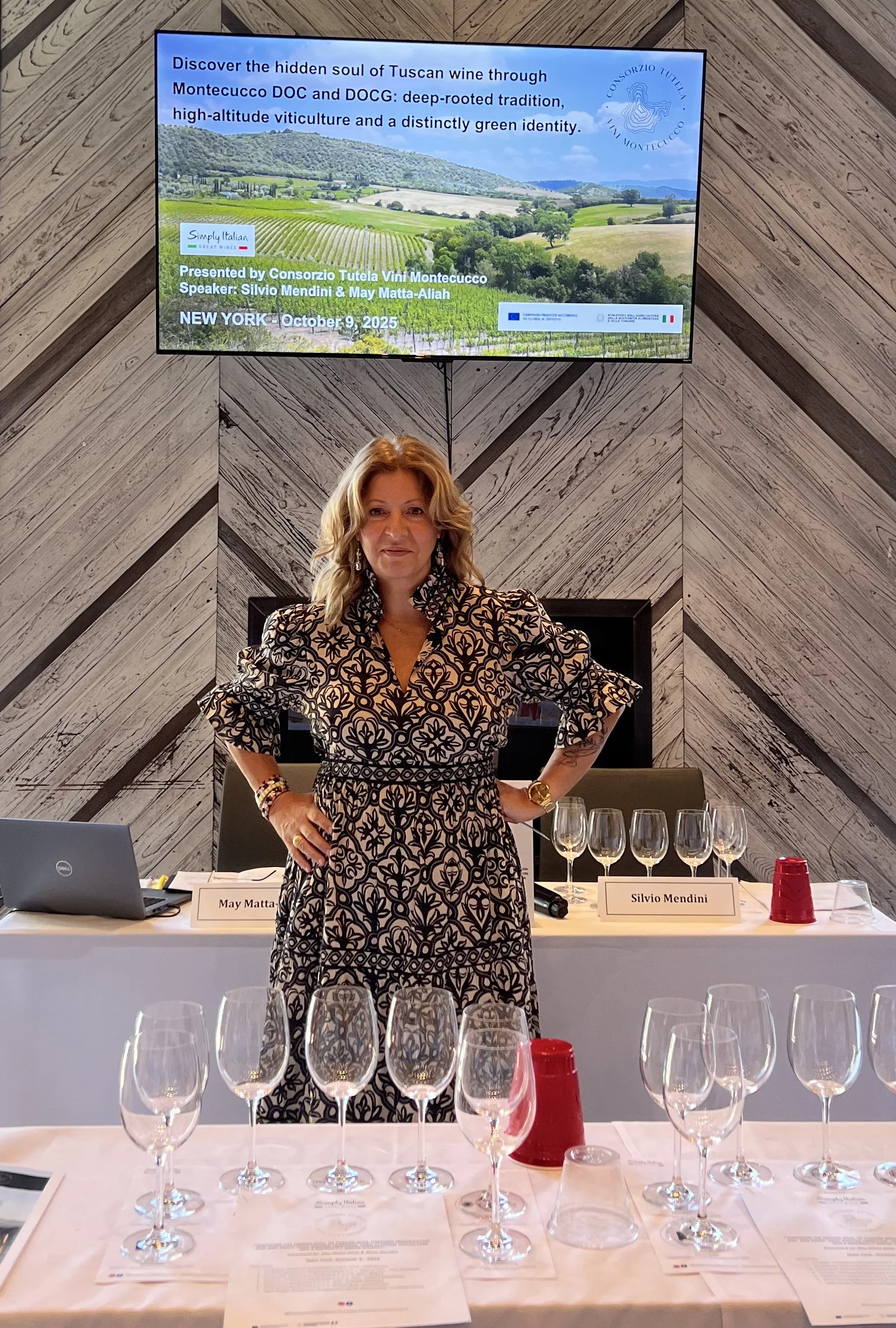 A woman standing behind a table with empty wine glasses at a wine tasting event. She is wearing a black and beige patterned dress. Behind her is a large screen displaying information about Tuscan wine, with a scenic vineyard image and event details.