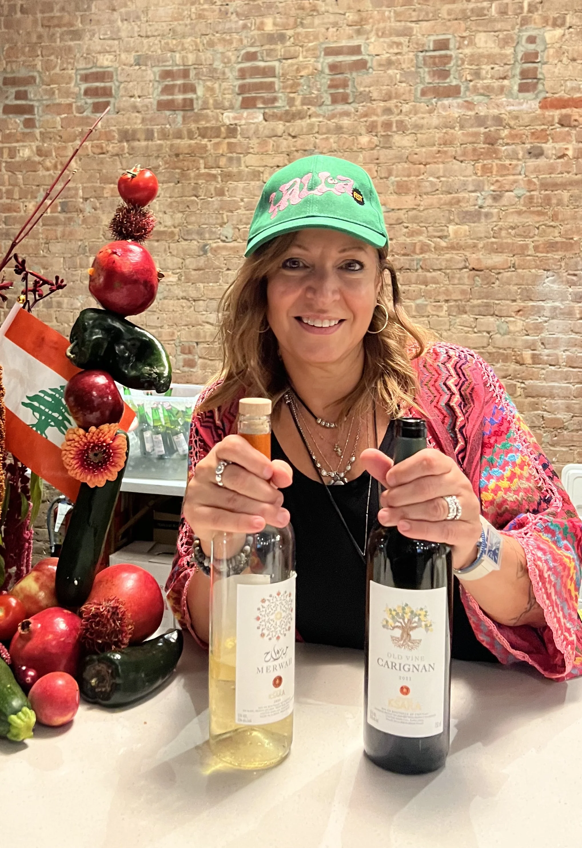 A woman smiling at a table with two bottles of wine, one white and one red, in front of her, with a decorative arrangement of apples, zucchinis, and a flower, and a brick wall in the background.