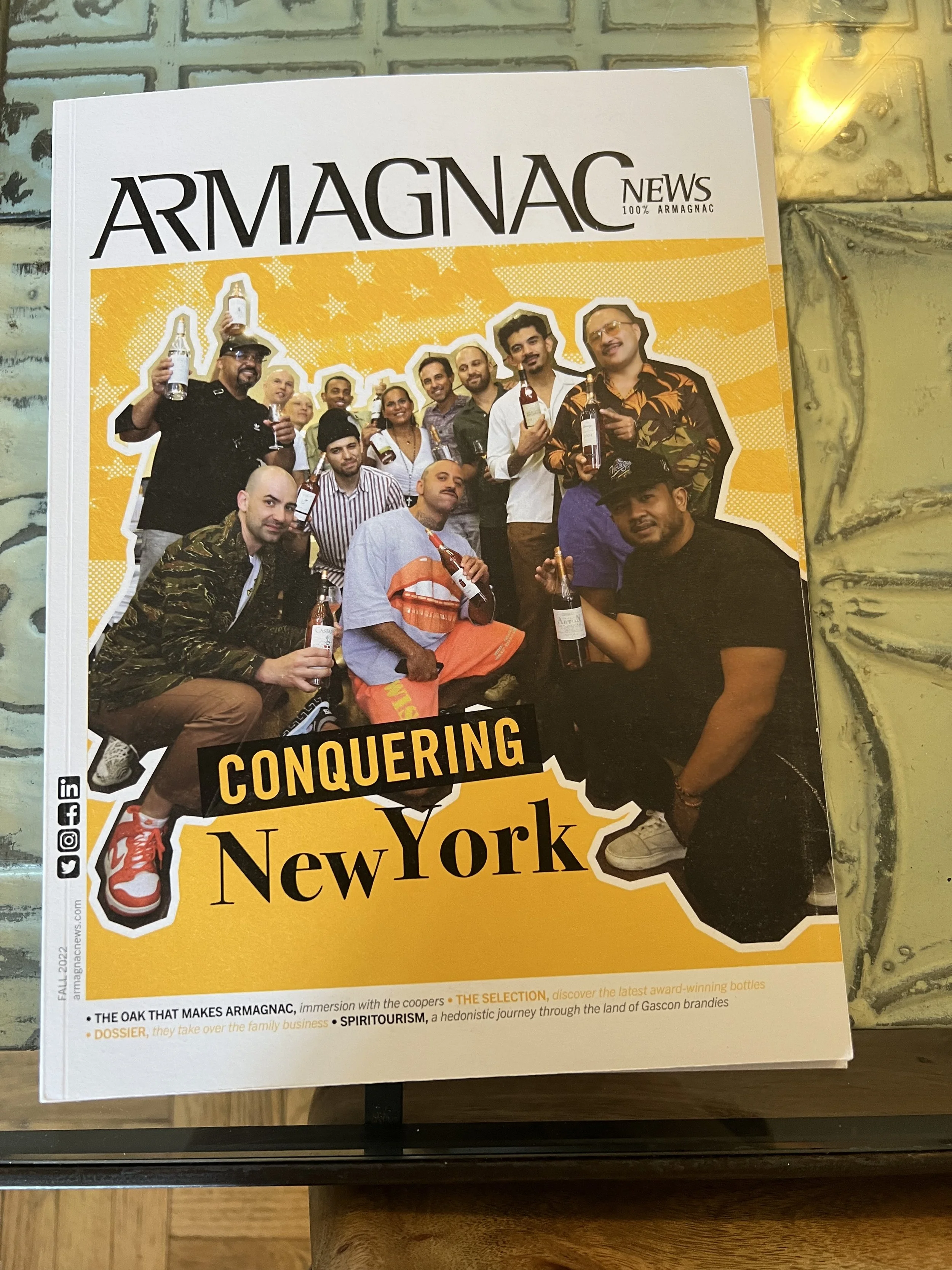 Cover of Armagnac News magazine featuring a group of people celebrating with bottles of Armagnac, with a yellow background and bold text 'Conquering New York'.