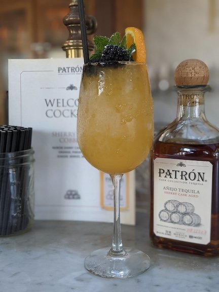 A yellow cocktail garnished with a lemon wedge, blackberries, and fresh mint leaves in a tall glass.