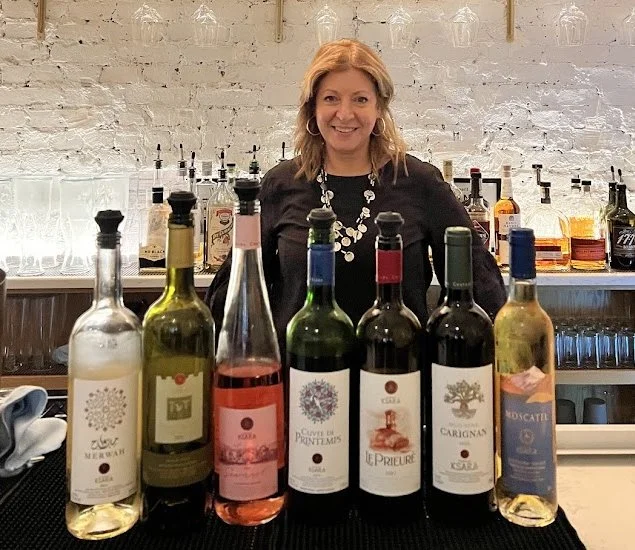 A woman standing behind a bar counter with eight bottles of wine in front of her, in a nicely lit bar with glasses hanging above.