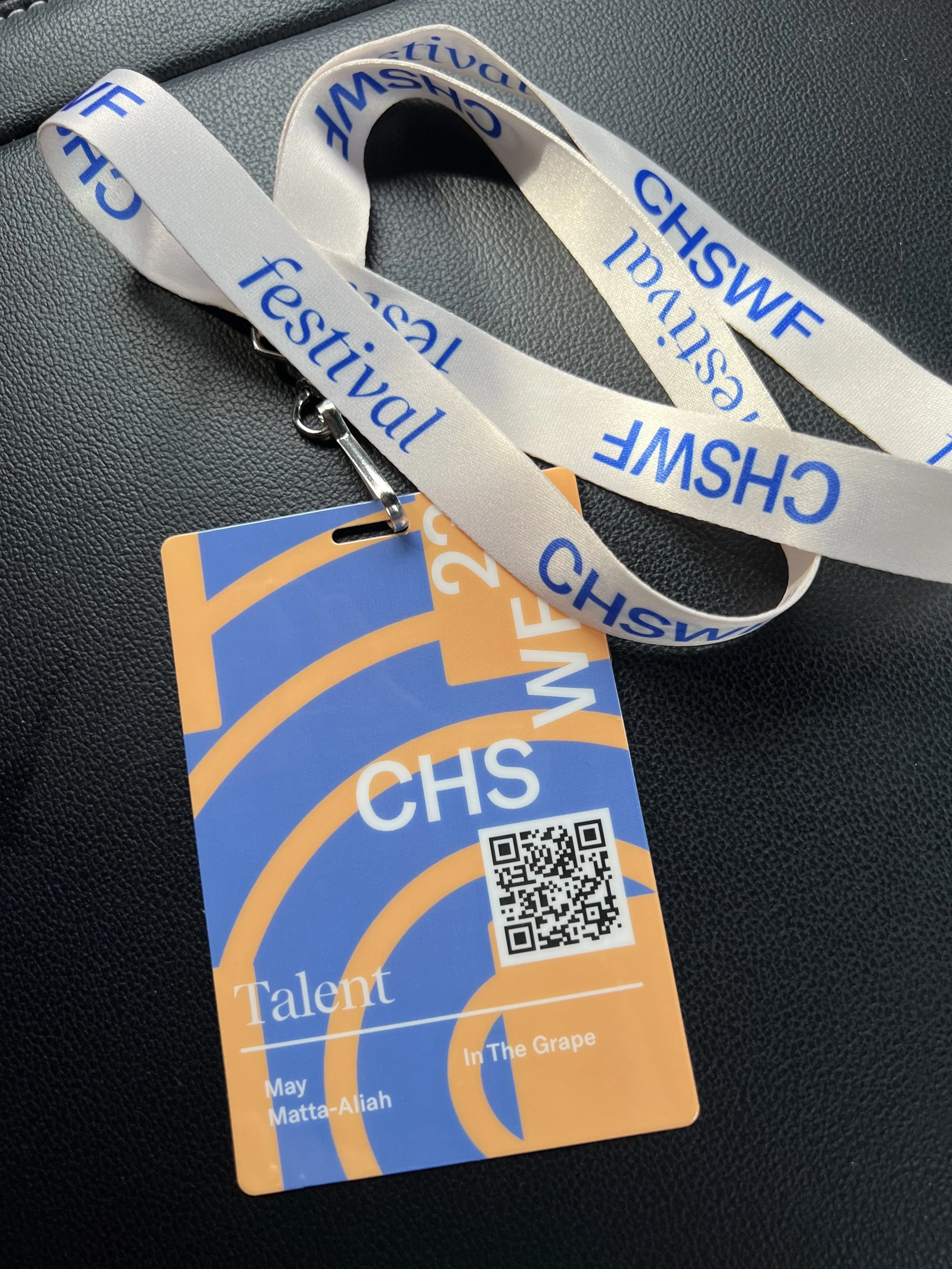 A white lanyard with blue text that reads 'CHSWF festival' and a badge with a QR code, the word 'Talent,' the name 'May Matta-Aliah,' and the phrase 'In The Grape.'