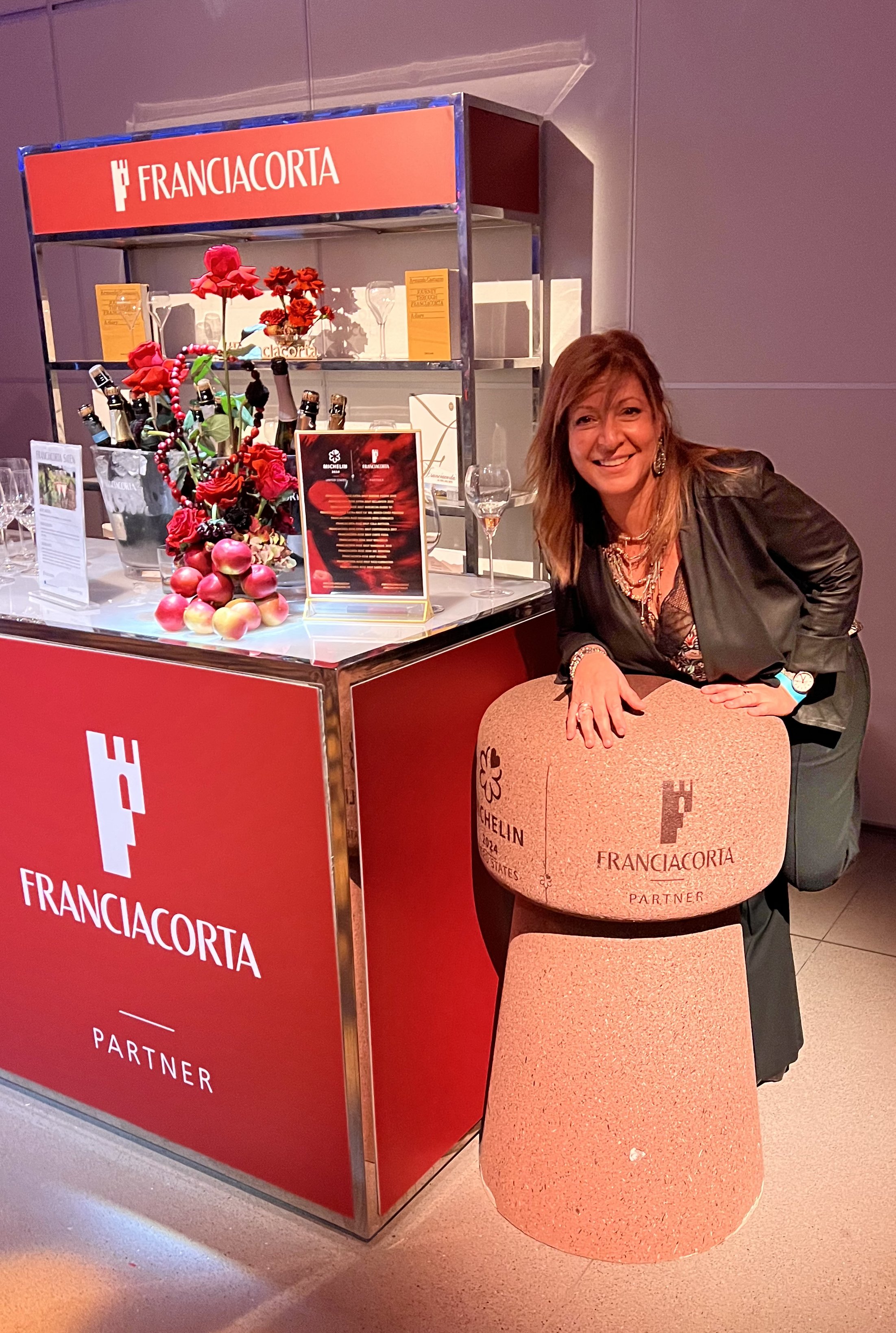A woman with light brown hair, wearing a black jacket, is smiling while leaning on a large beige stone furniture piece with 'FRANCIACORTA' written on it, next to a red display stand labeled 'FRANCIACORTA PARTNER'. The stand has a floral arrangement w