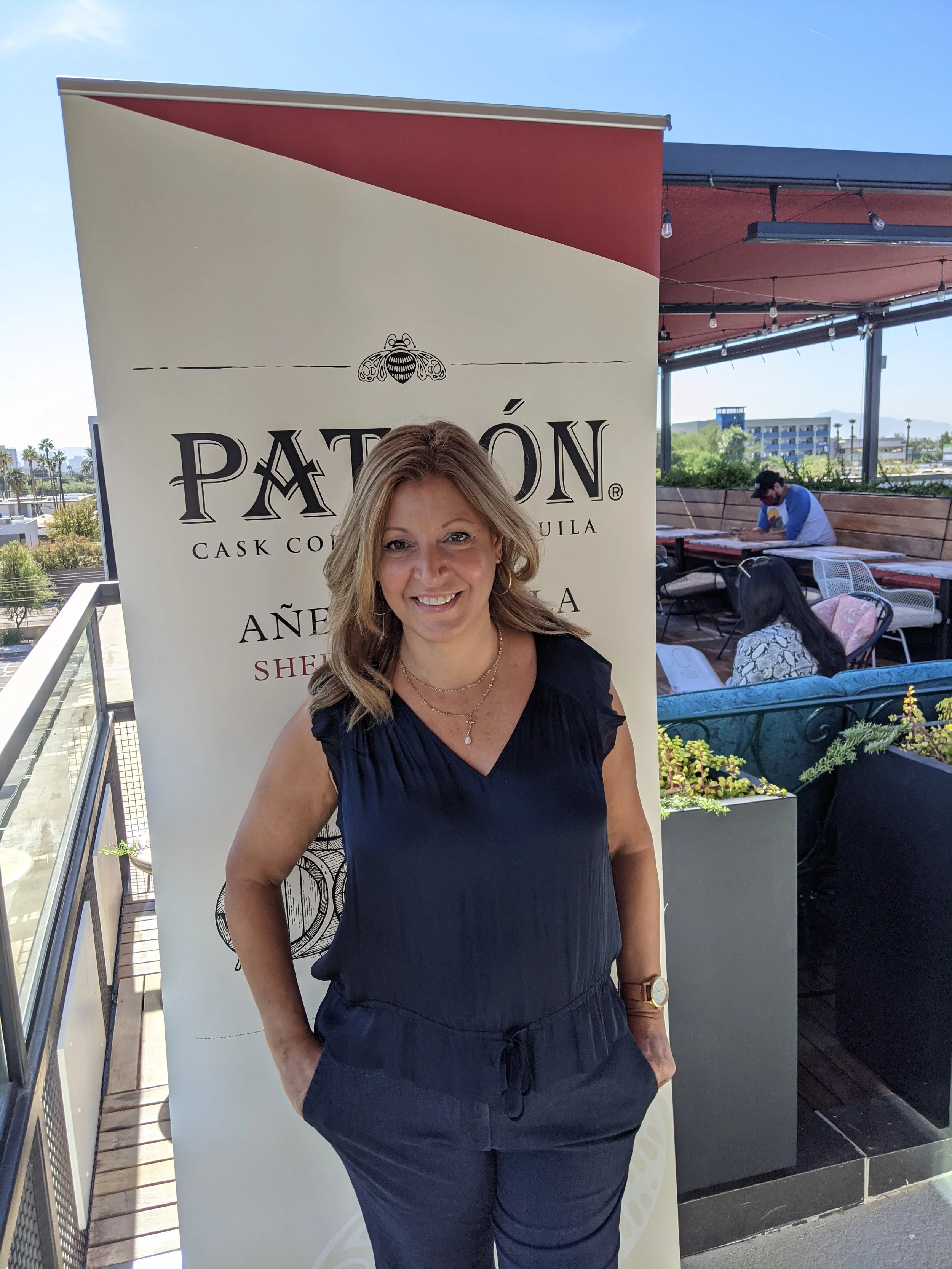 Woman standing in front of a Patagonia Tequila banner on a rooftop patio with other people seated at tables, clear blue sky in the background.