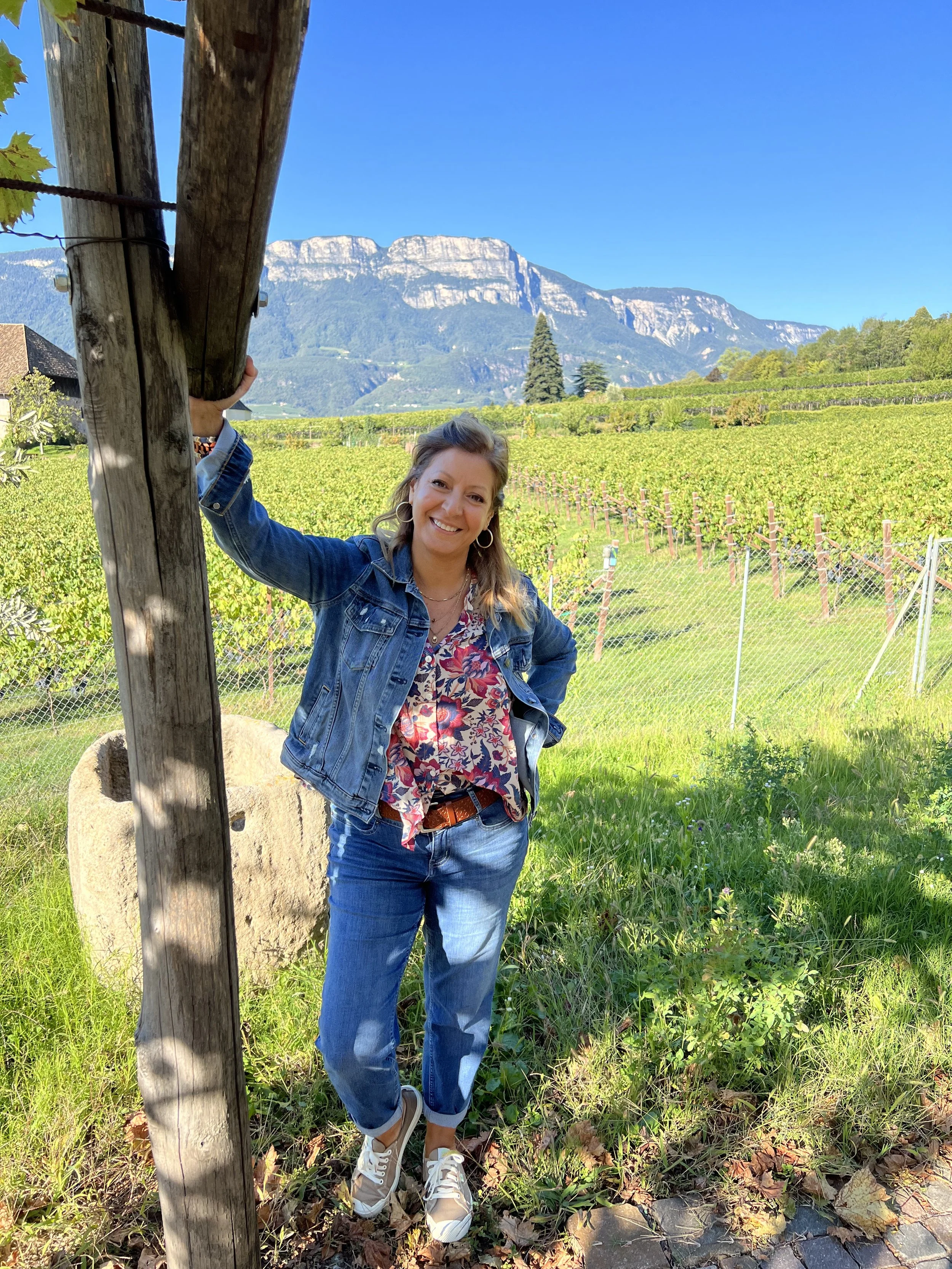 Woman in denim jacket and jeans smiling outdoors in a vineyard, with mountains in the background.
