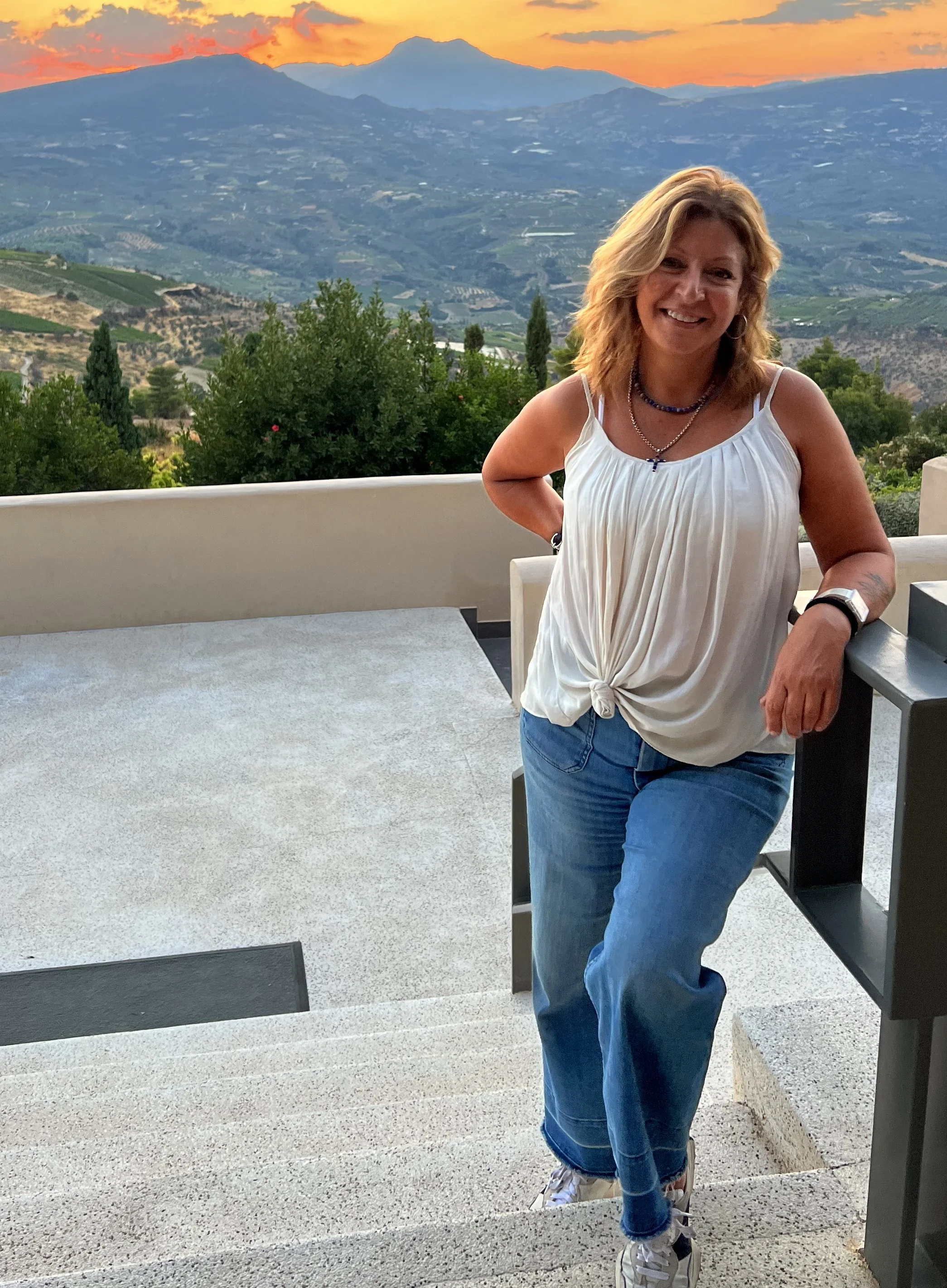 A woman leaning on a staircase railing outdoors during sunset, with mountains and a sunset sky in the background.