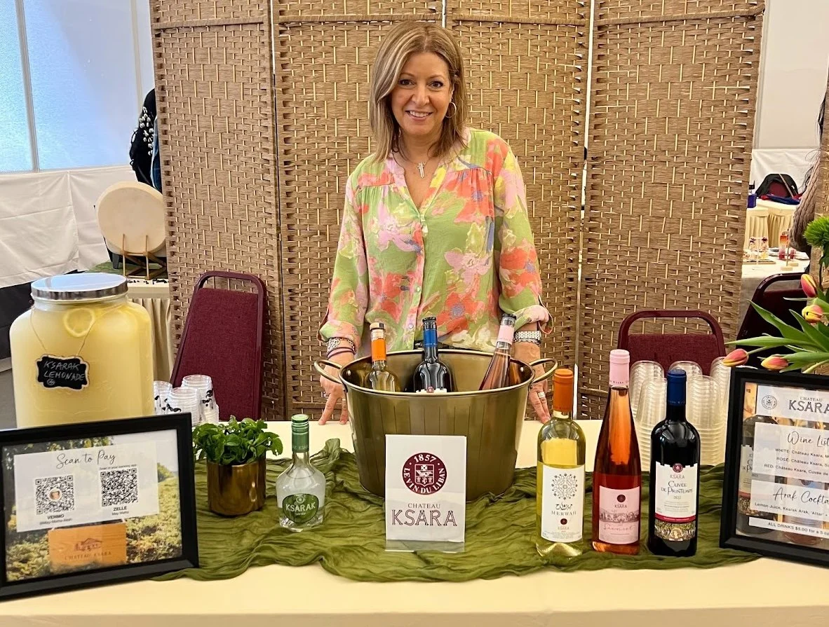 A woman standing behind a table with bottles of wine, a small potted plant, and signs advertising wine and lemonades at a wine tasting event.