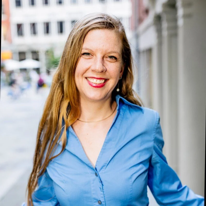A woman with long red hair, smiling with teeth showing, wearing a blue button-up shirt and pearl earrings, standing outdoors in an urban setting.
