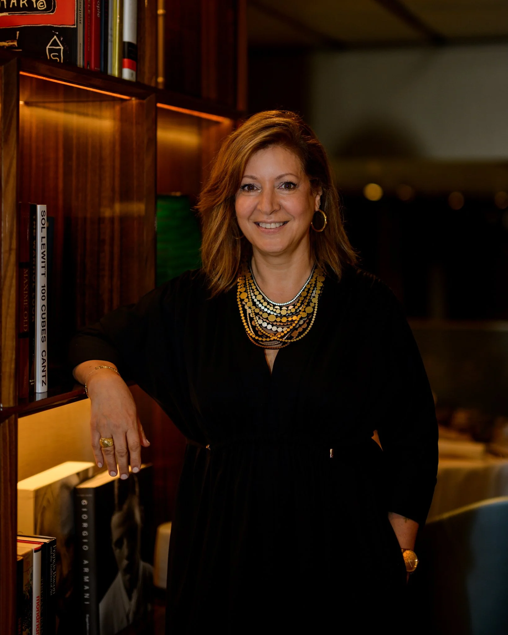 A woman with shoulder-length light brown hair and hoop earrings, smiling and wearing a black dress with layered gold and silver necklaces, standing in front of a wooden bookshelf with books.