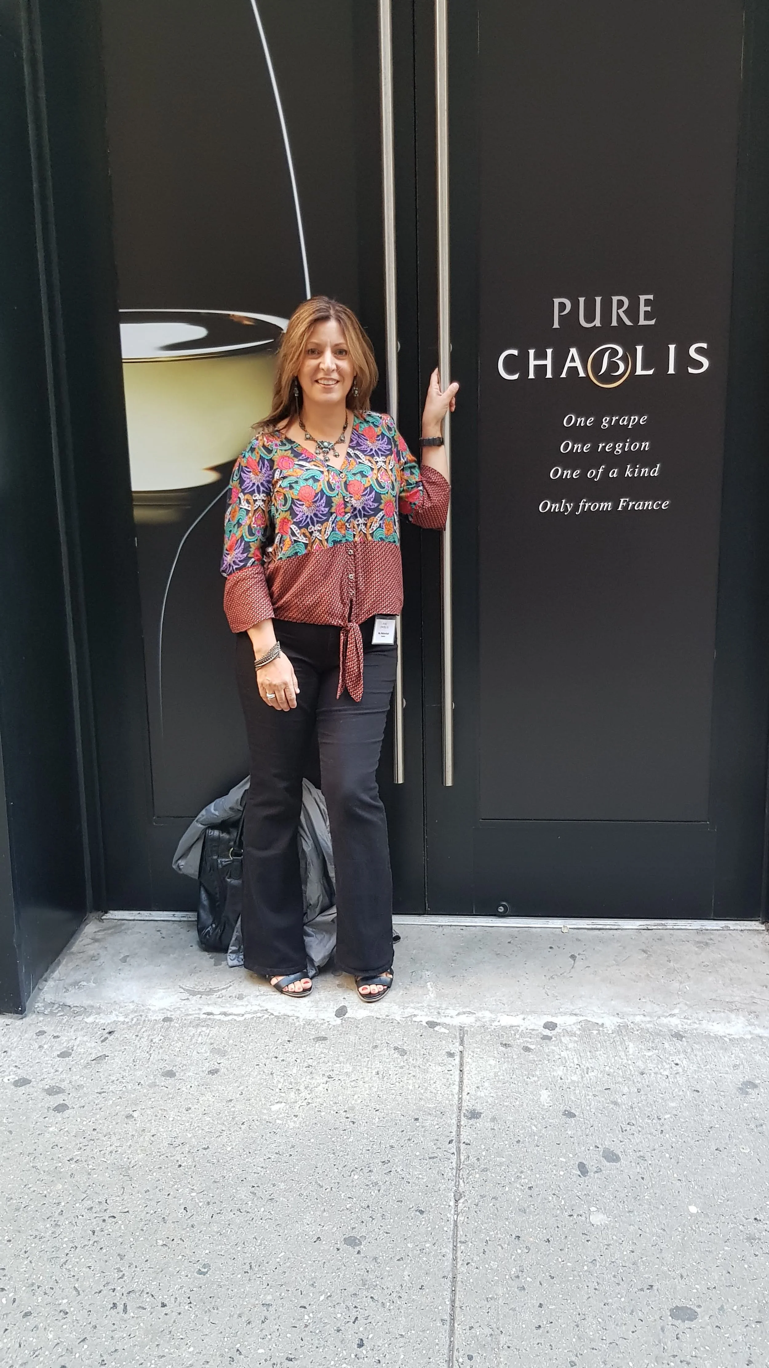 Woman standing in front of a large black door with a wine image and text that reads 'Pure Chablis - One grape, One region, One of a kind, Only from France.' She is smiling, wearing a colorful blouse with floral patterns, black pants, and open-toed bl