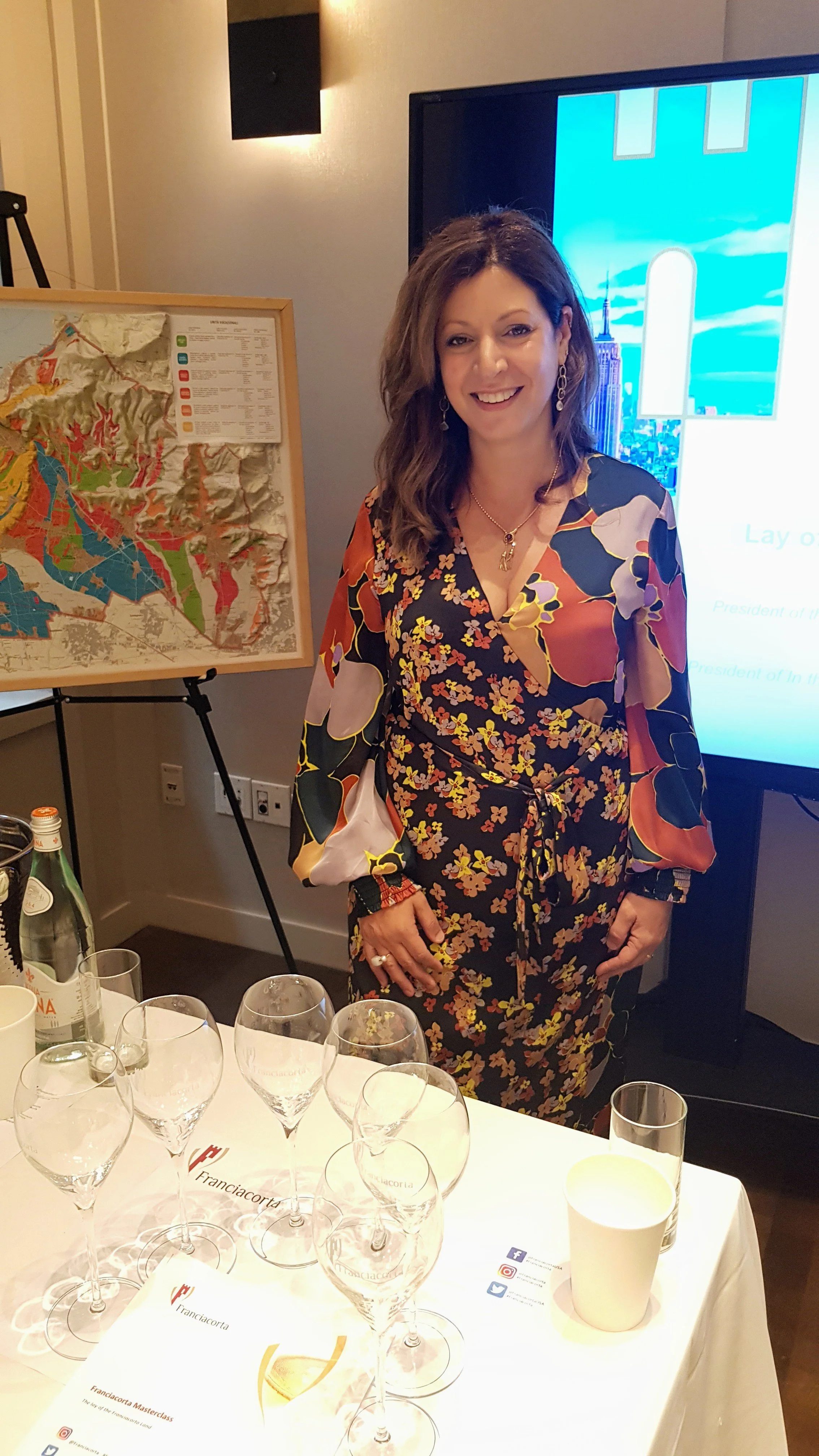 A woman standing at a table with multiple empty wine glasses, a water bottle, and a paper cup. She is smiling and wearing a colorful floral dress. Behind her, there is a large screen with a presentation slide and a map on an easel.