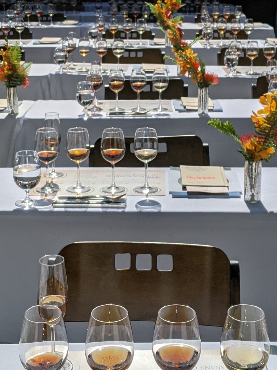 Several rows of tables set for a tasting event, each with multiple glasses of wine, floral centerpieces in vases, and printed menus.