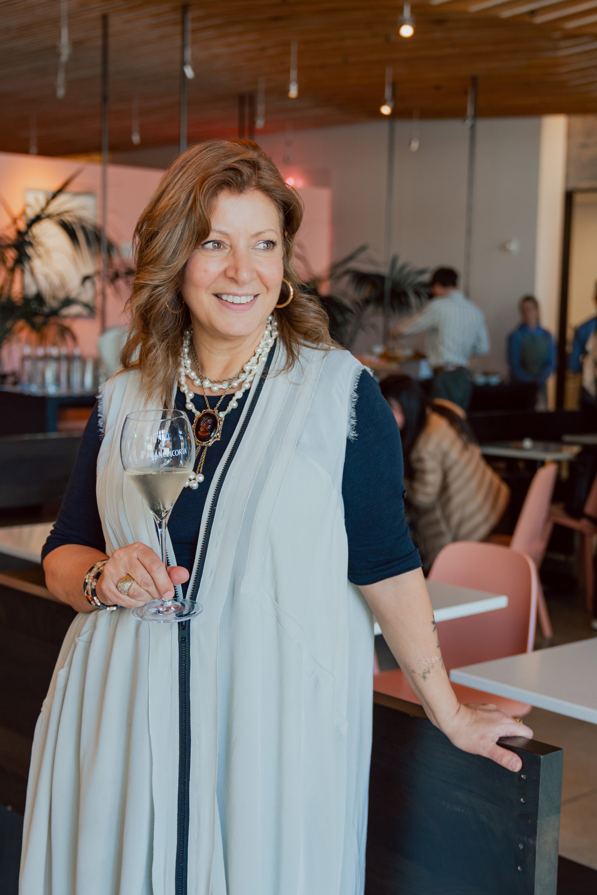 A woman holding a glass of white wine in a restaurant or cafe with other people in the background.