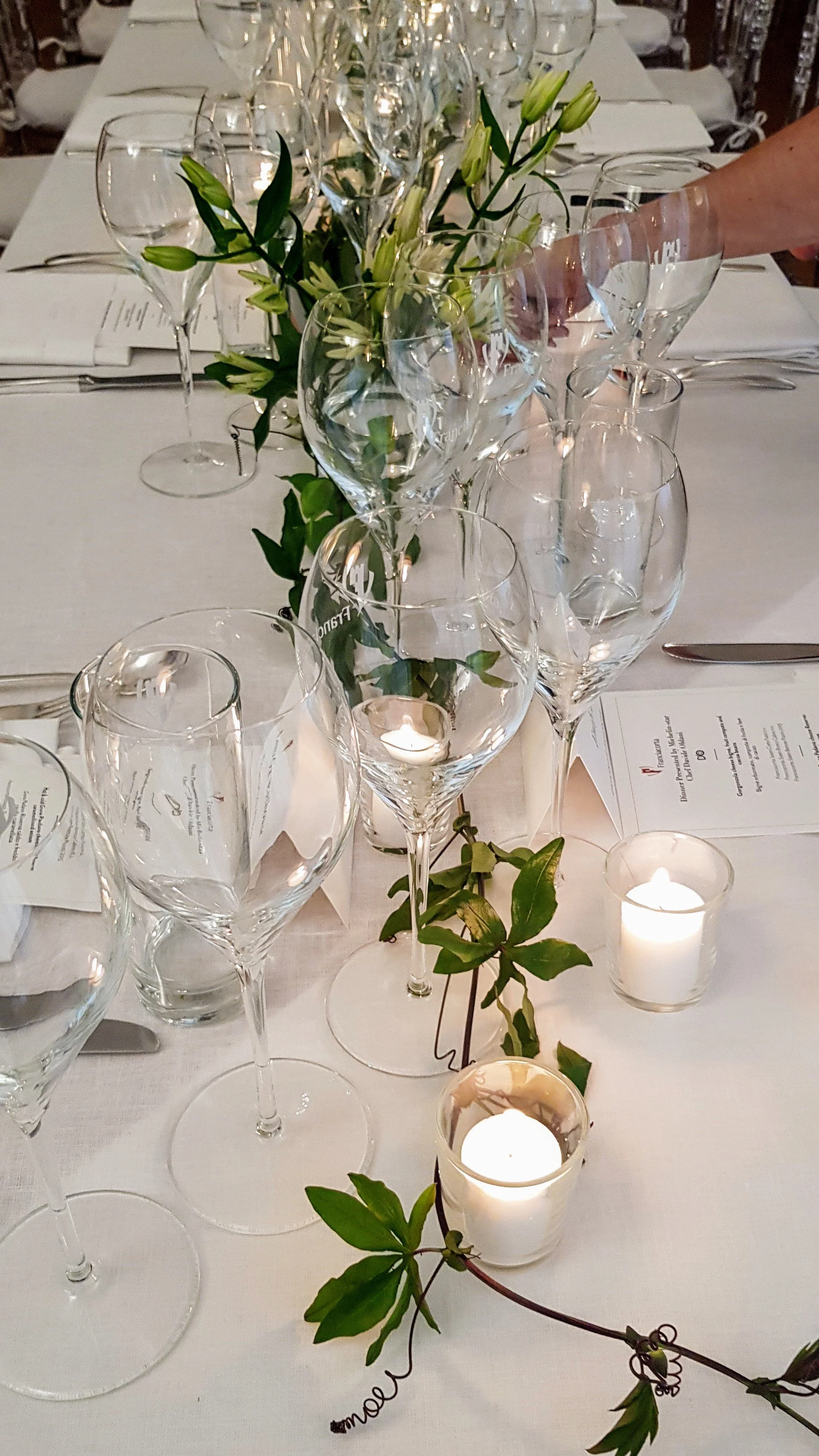 A table set for a formal event with empty wine glasses, a floral centerpiece with white lilies, lit candles, and place settings on a white tablecloth.