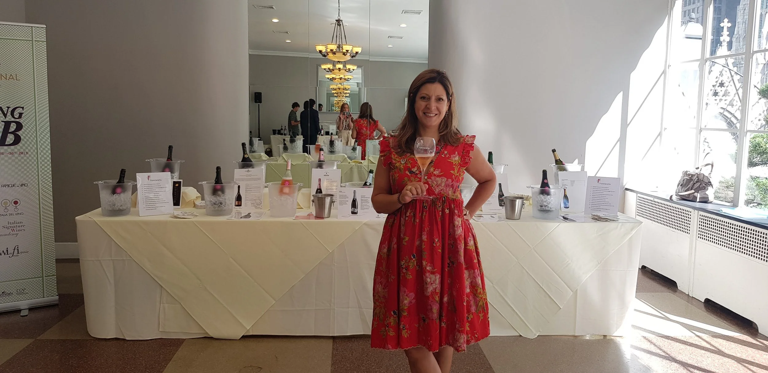 Woman in red floral dress holding a wine glass at a wine tasting event with bottles and informational cards on a table behind her.