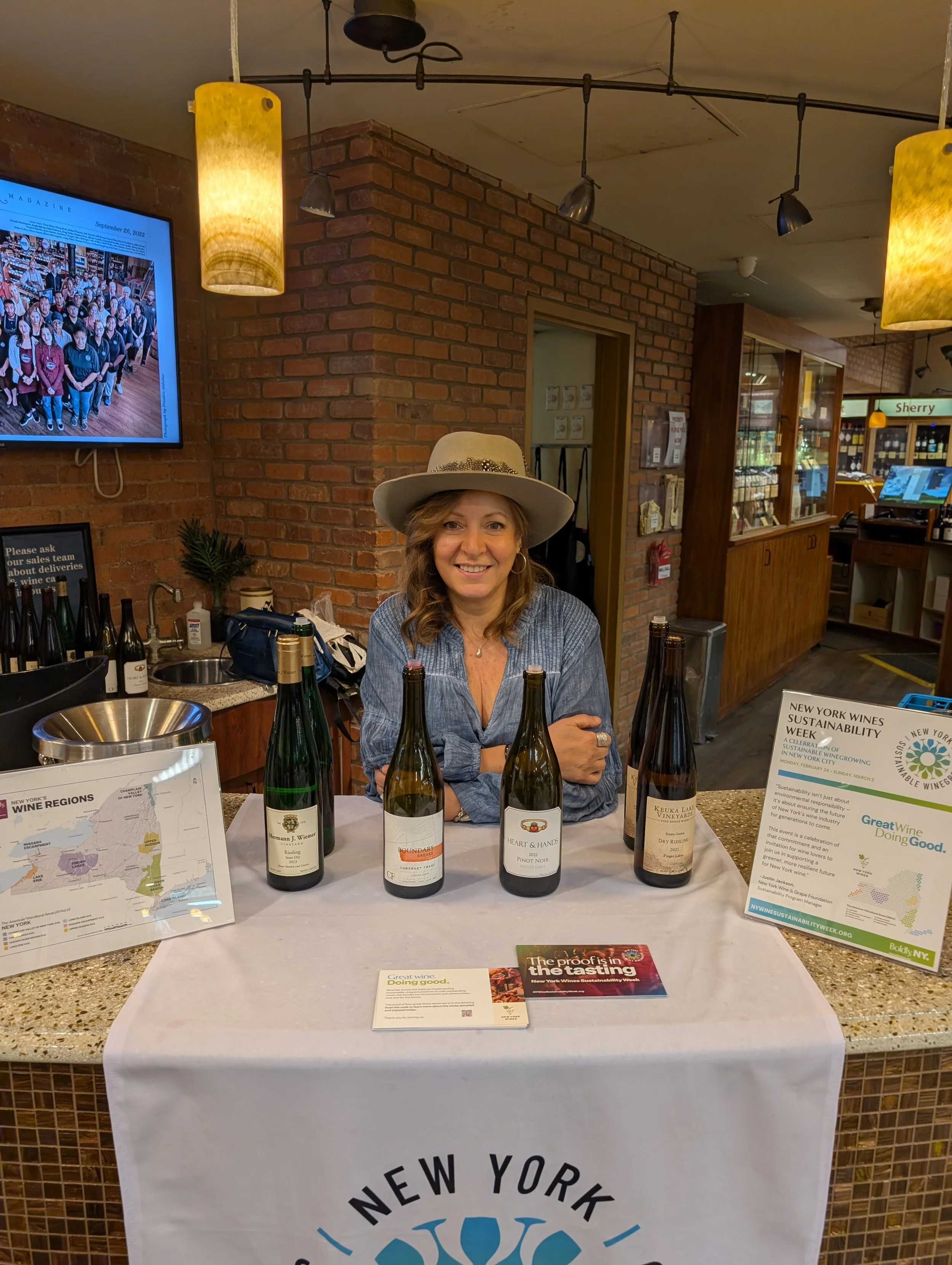 A woman wearing a wide-brimmed hat and denim shirt standing behind a table with bottles of wine, promotional materials, and informational signs about New York wines and sustainability in wine production.