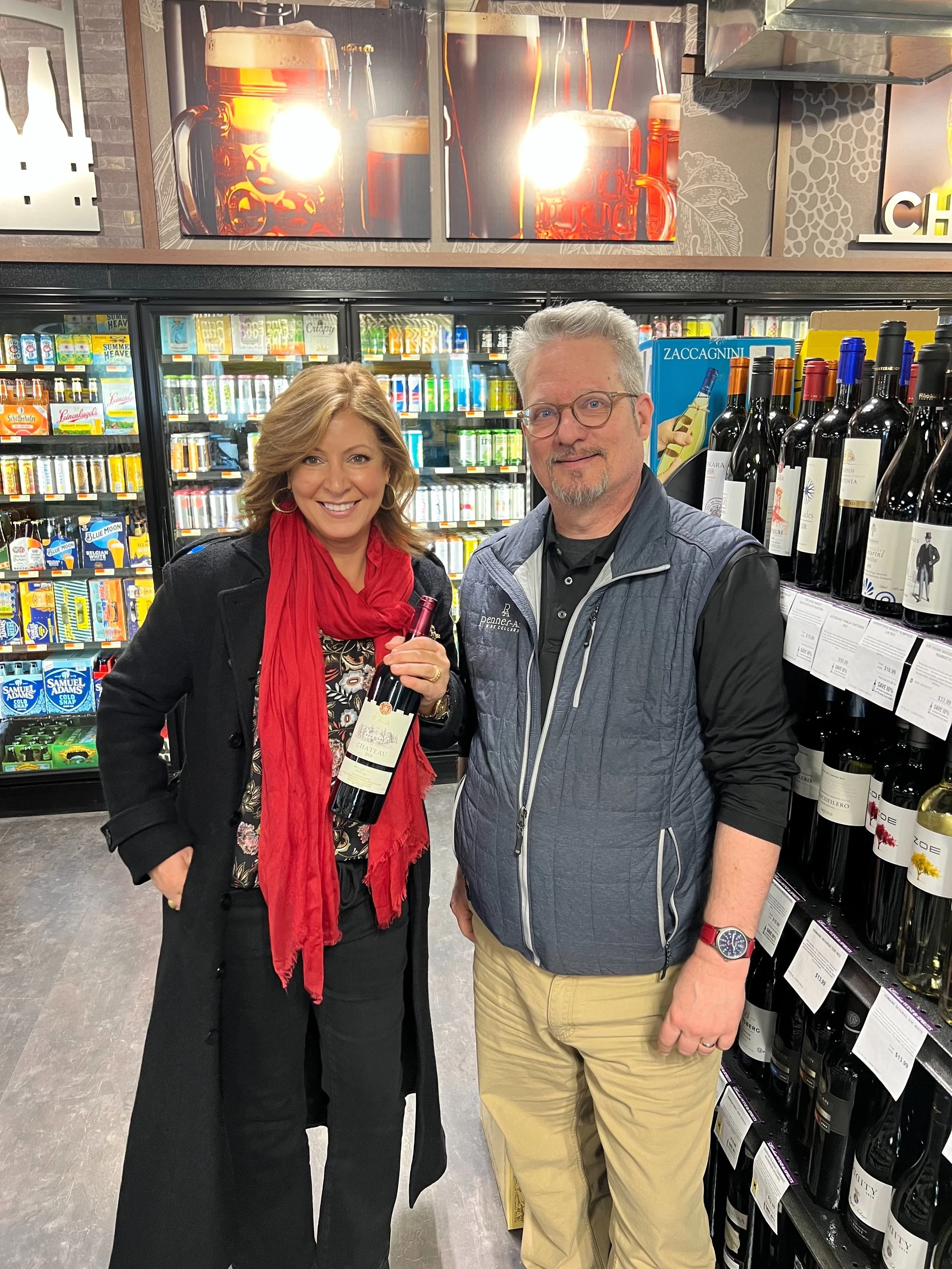 A woman and a man standing in a grocery store aisle, smiling. The woman is holding a bottle of red wine, and the man is wearing glasses and a vest. There are wine bottles on the shelves behind them.