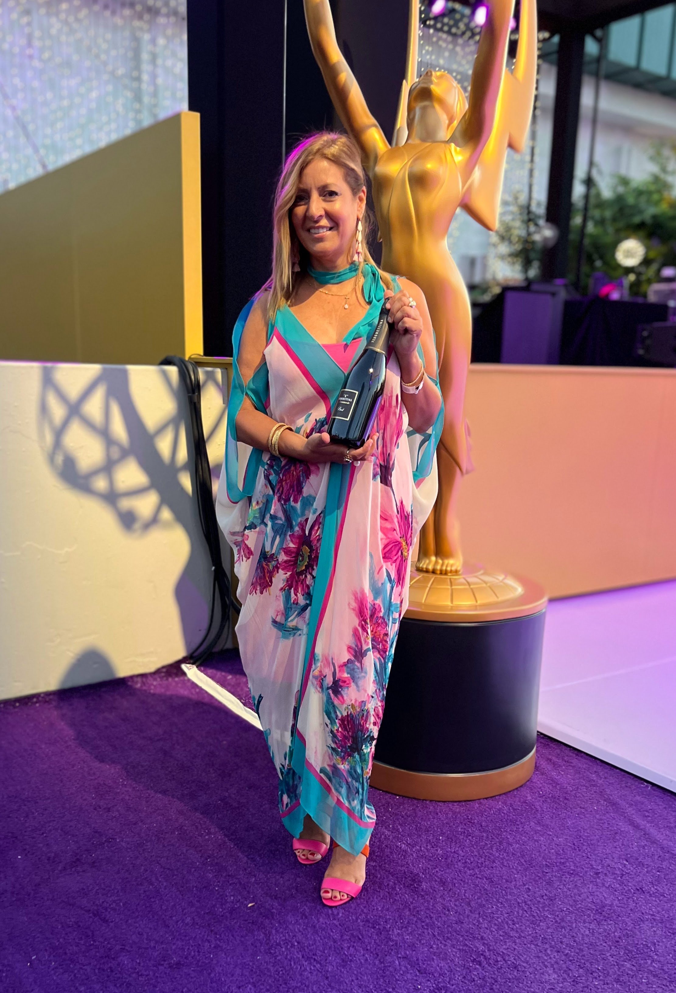 A woman in a colorful floral dress and pink open-toe shoes holding a bottle of champagne, standing on purple carpet in front of a golden statue, smiling at the camera.