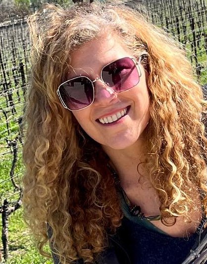Woman with curly blonde hair wearing pink sunglasses smiling outdoors in front of a black fence.