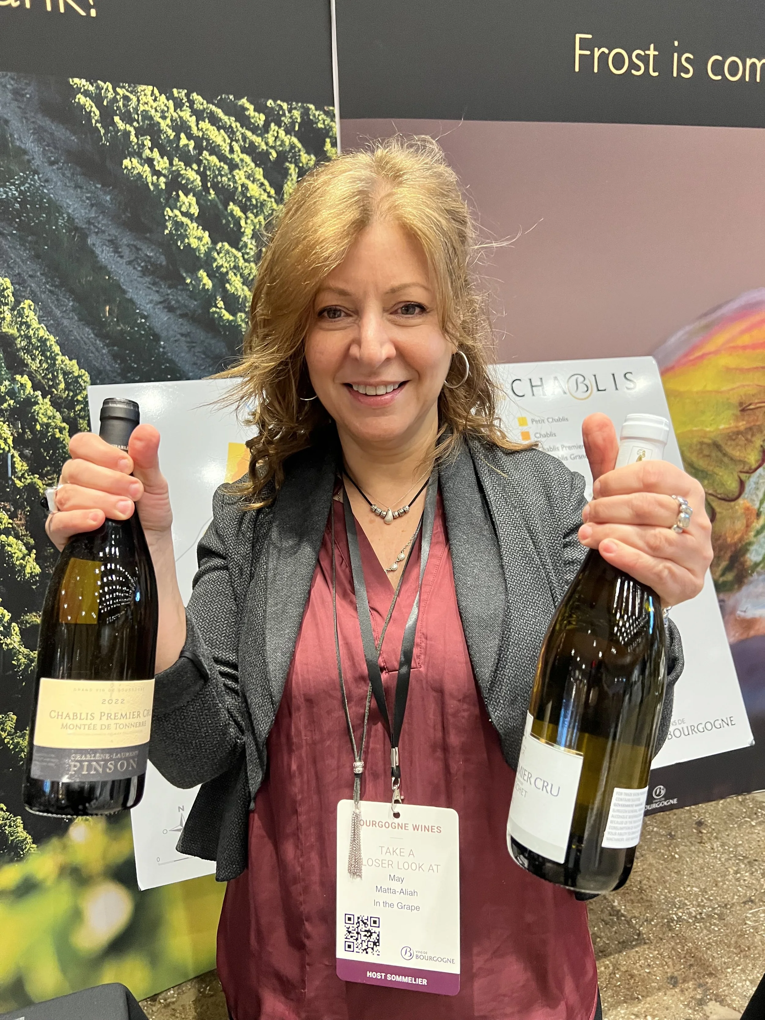 A woman smiling and holding two bottles of wine, wearing a gray blazer and a red top, with a name badge hanging around her neck, standing in front of a wall display advertising wines from Burgundy.