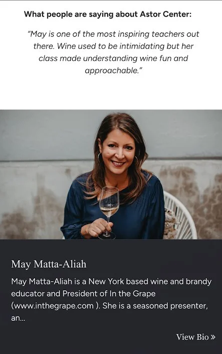 A woman with long brown hair, wearing a blue blouse, smiling, sitting indoors, holding a glass of white wine.
