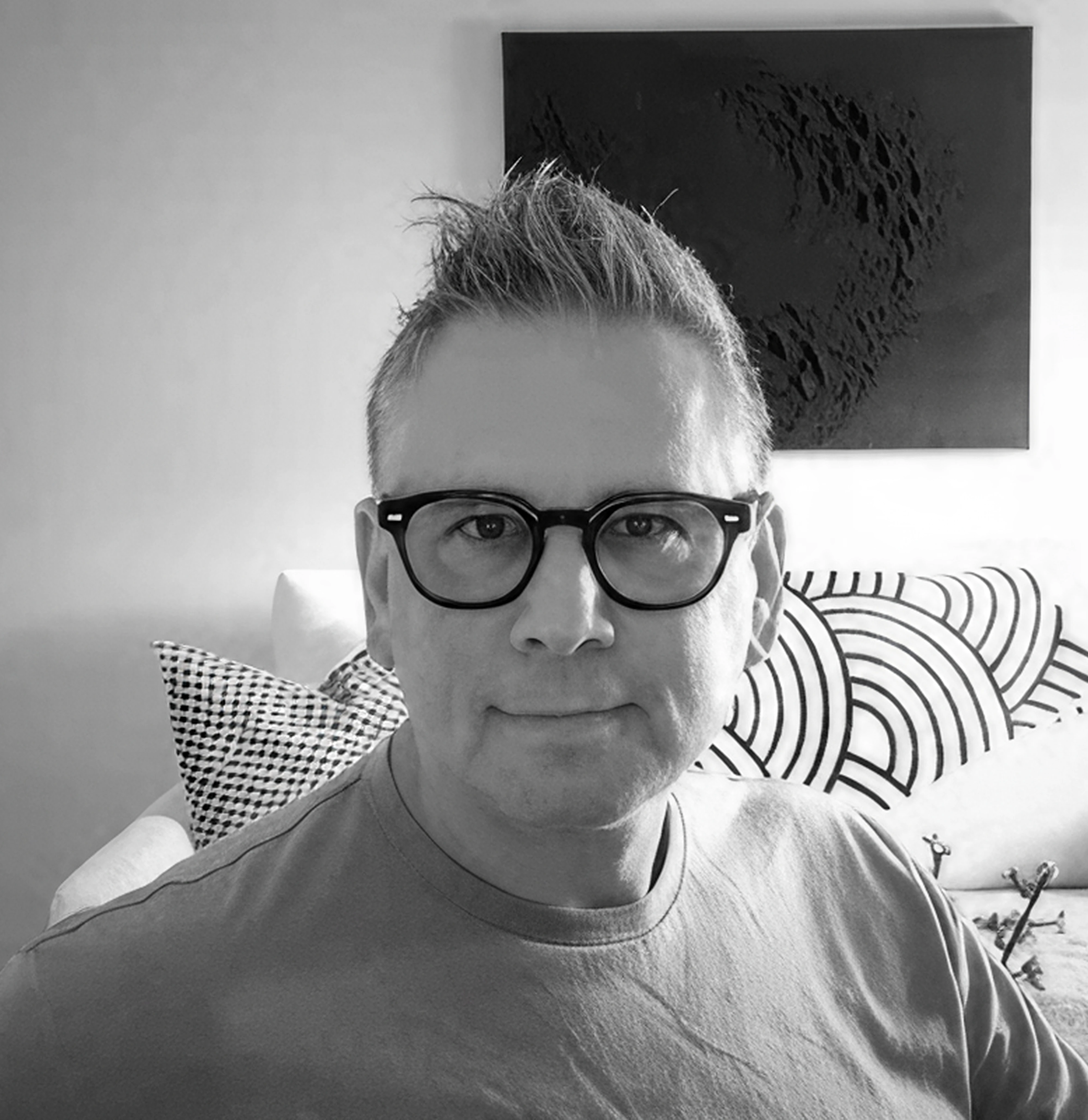 Black and white photo of a man wearing glasses, sitting on a sofa with patterned pillows and artwork hanging on the wall in the background.