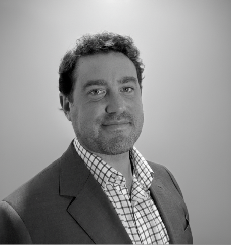 Black and white photo of a man with curly hair wearing a suit and checkered shirt, looking at the camera with a slight smile.