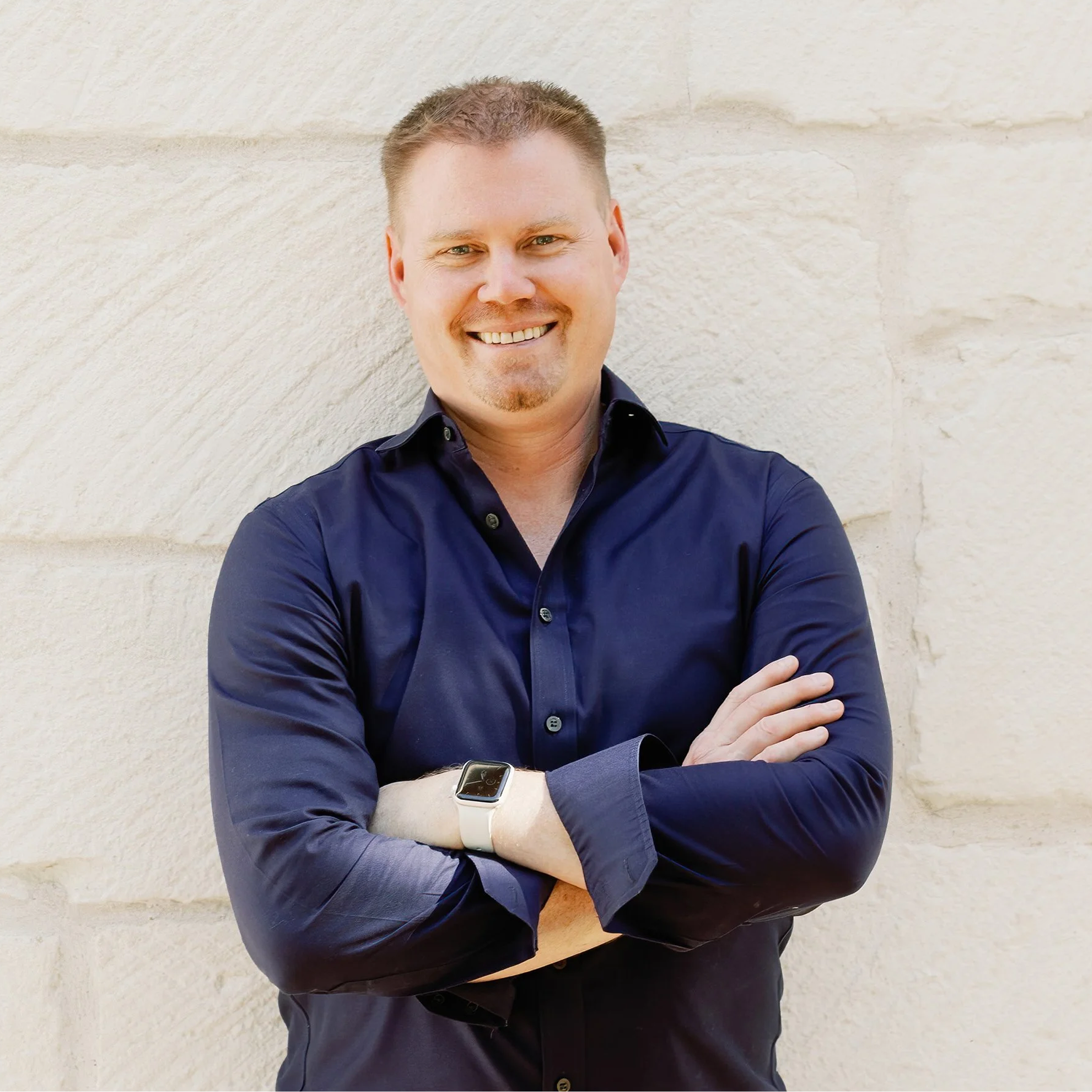 A smiling man in a navy blue shirt with rolled-up sleeves stands with arms crossed in front of a beige stone wall.