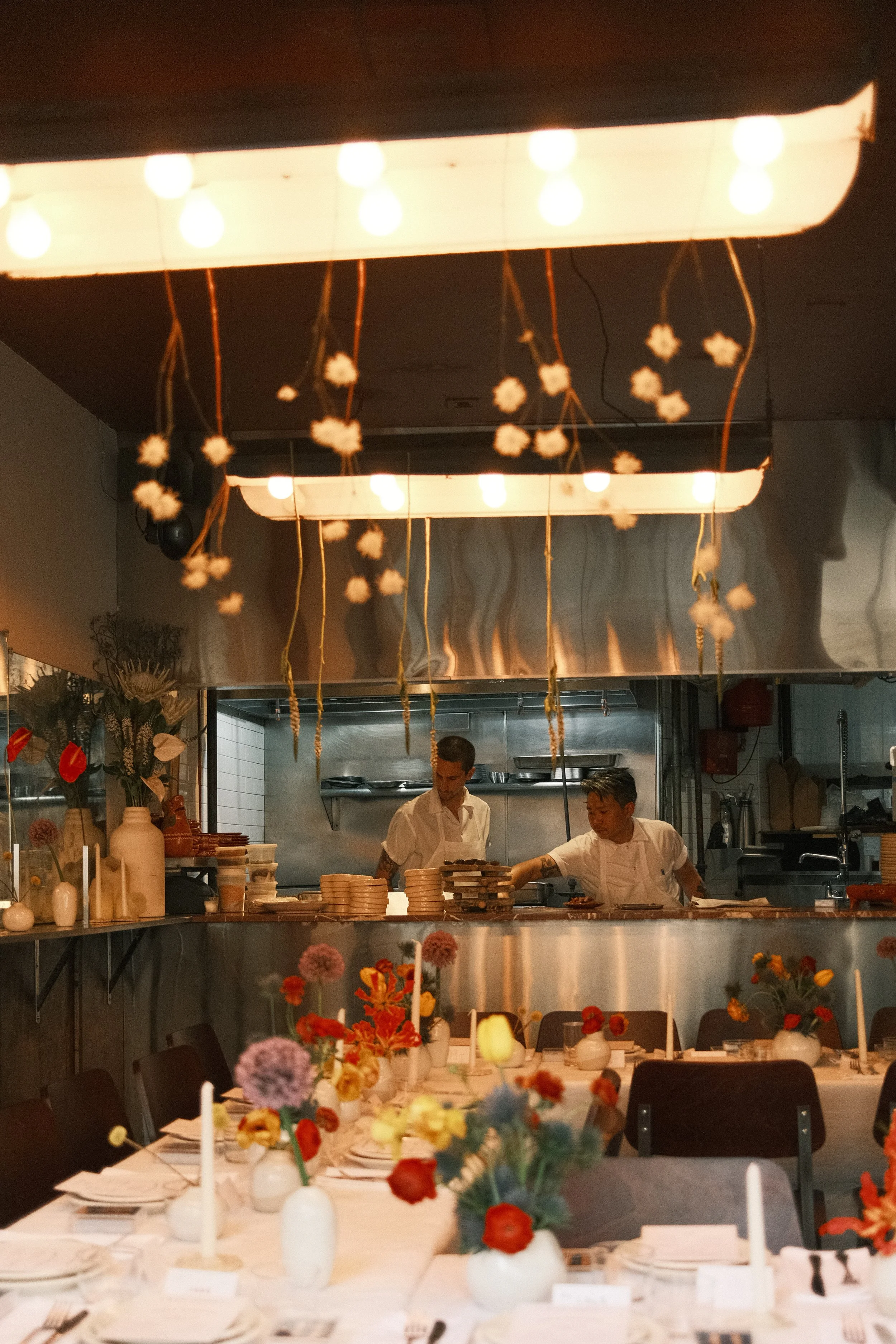 Interior of a restaurant or event space with a long decorated table set with plates, cutlery, candles, and floral arrangements, and two chefs working in an open kitchen area above.