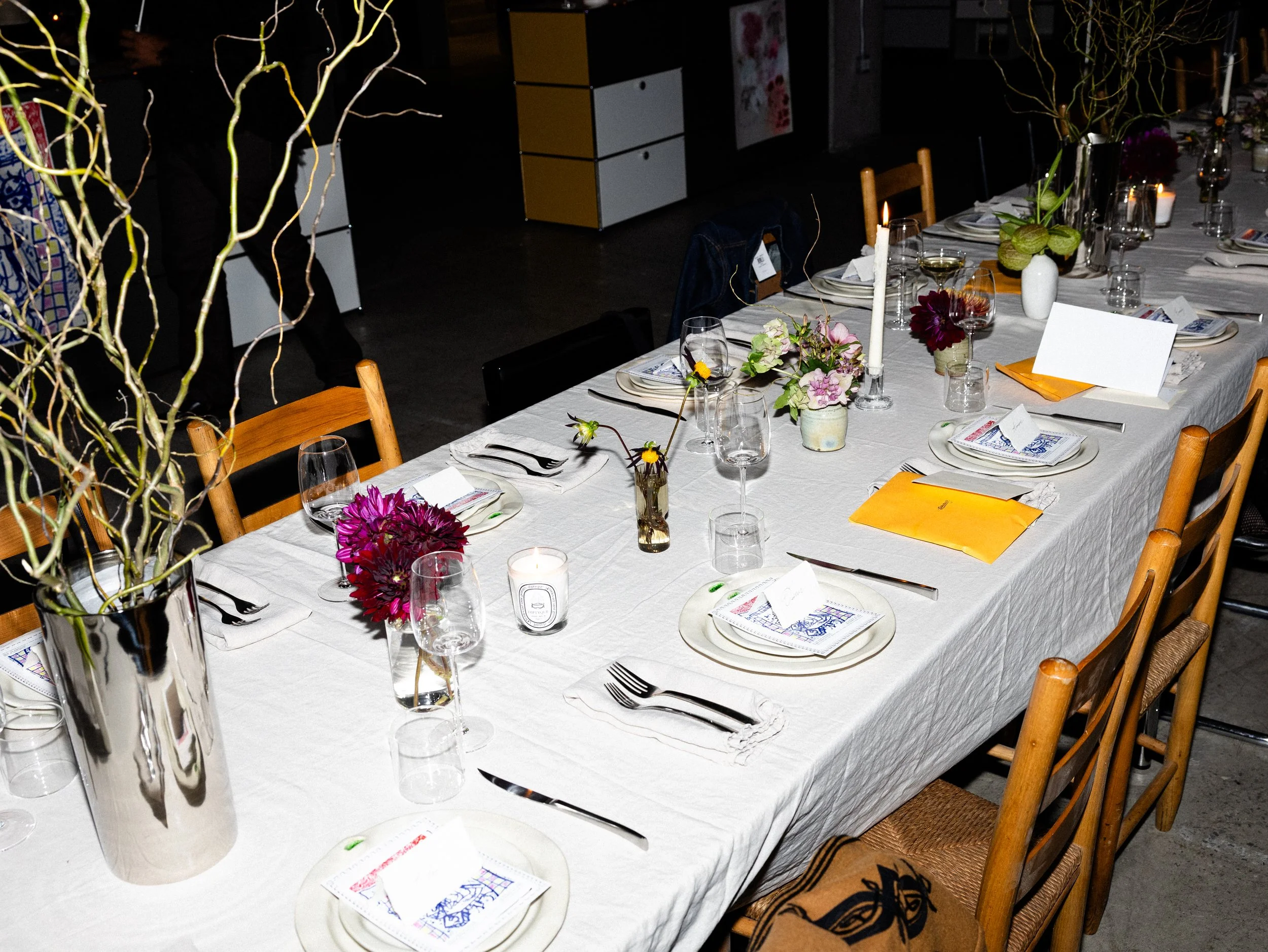 A long dining table set for a formal meal, decorated with flower arrangements, candles, white tablecloths, folded napkins, and numerous wine glasses. There are chairs around the table and a dark background with some cabinets and decorations.