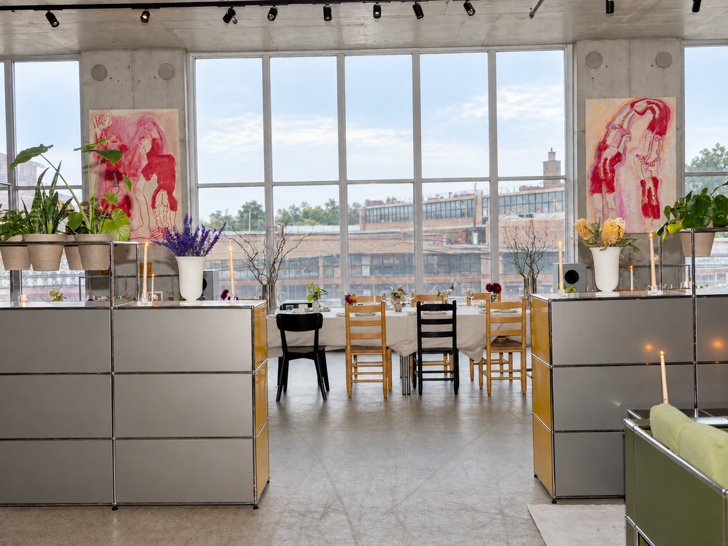 Interior of a modern dining space with a long table, colorful chairs, floral arrangements, candles, and large windows overlooking a cityscape.