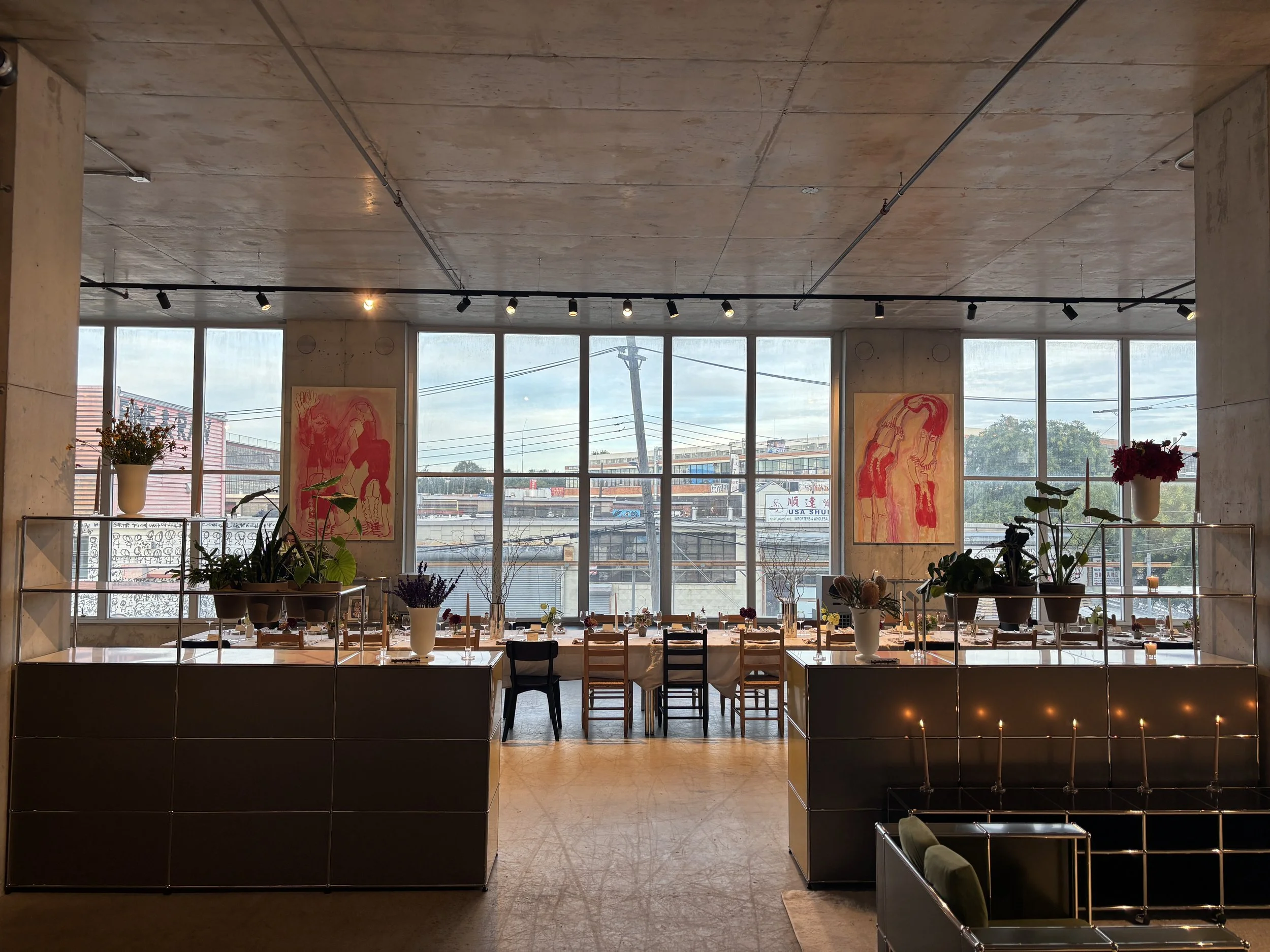 Interior of an event with large windows, plants, long dinner table, plates, glassware, artwork on the walls, tables decorated with flowers, and ceiling-mounted spotlights.
