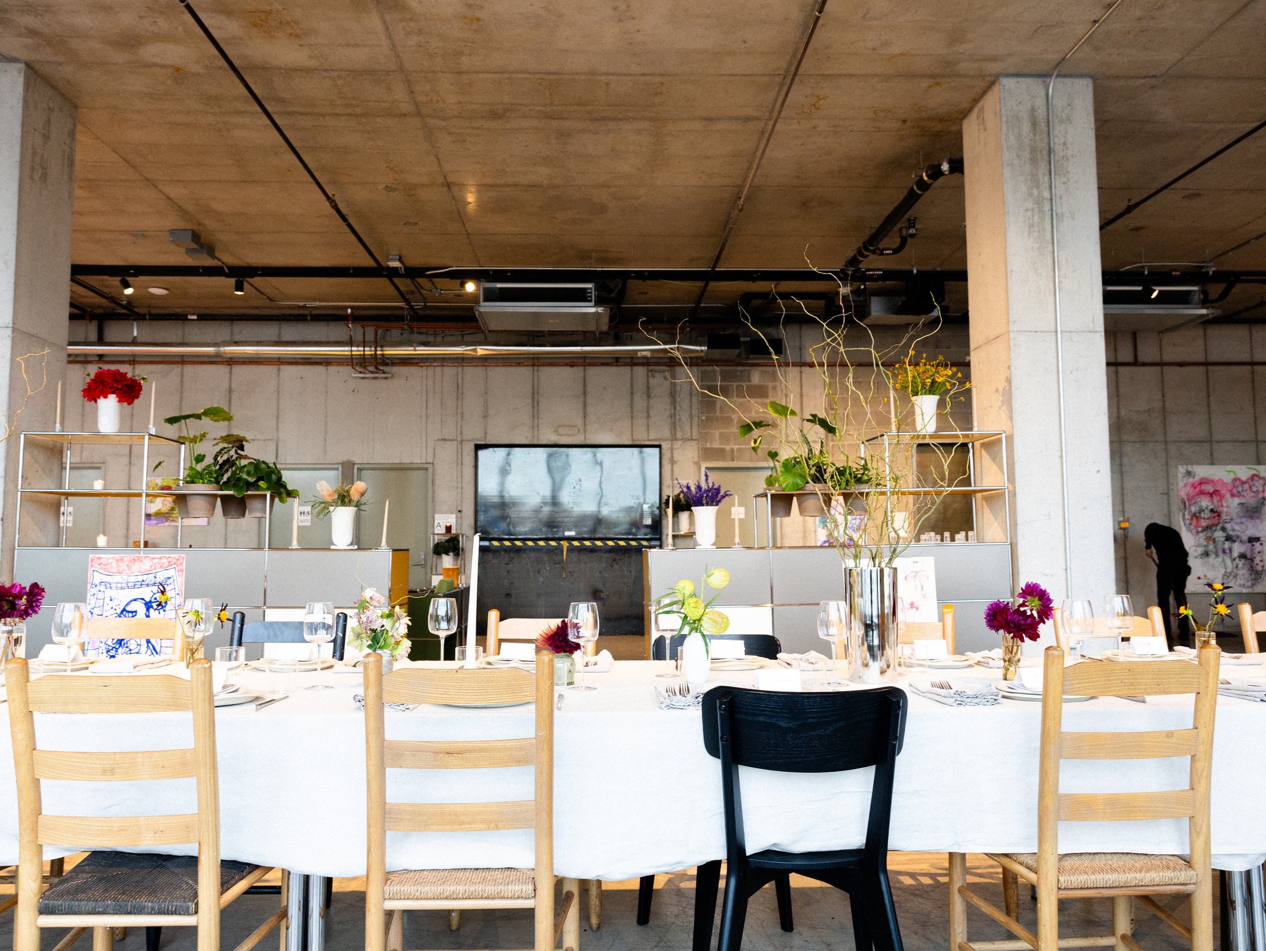 A decorated event space with a long table set for a meal, decorated with flowers. The background has an industrial style with visible pipes and concrete walls.