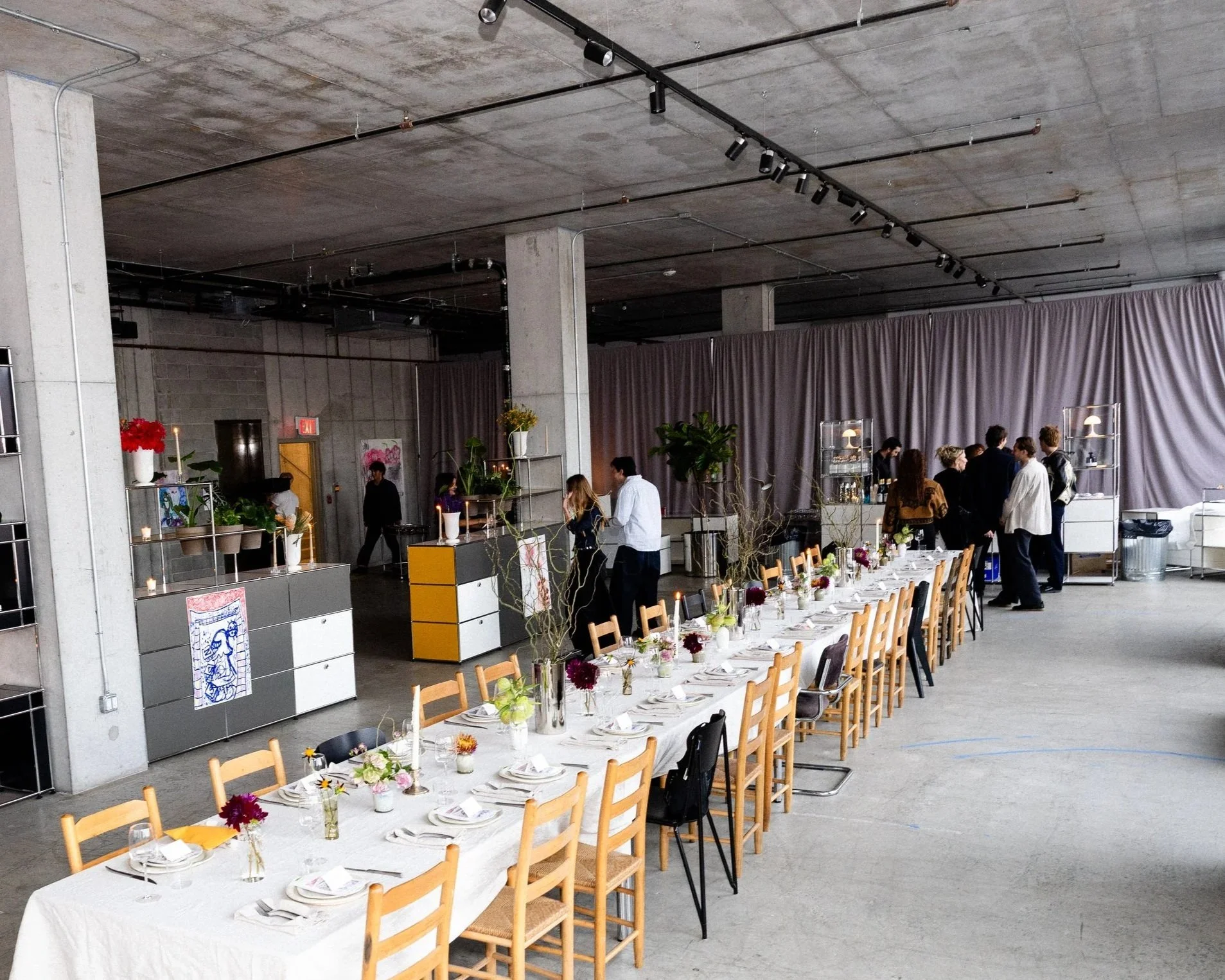 Long rectangular dinner table with a white tablecloth, set with plates, glasses, napkins, and small flower arrangements in vases. Wooden chairs are arranged along the table. In the background, people are gathered near a gray curtain, and the room has an industrial appearance with concrete walls and ceiling, exposed beams, track lighting, and some potted plants and artwork for decoration.