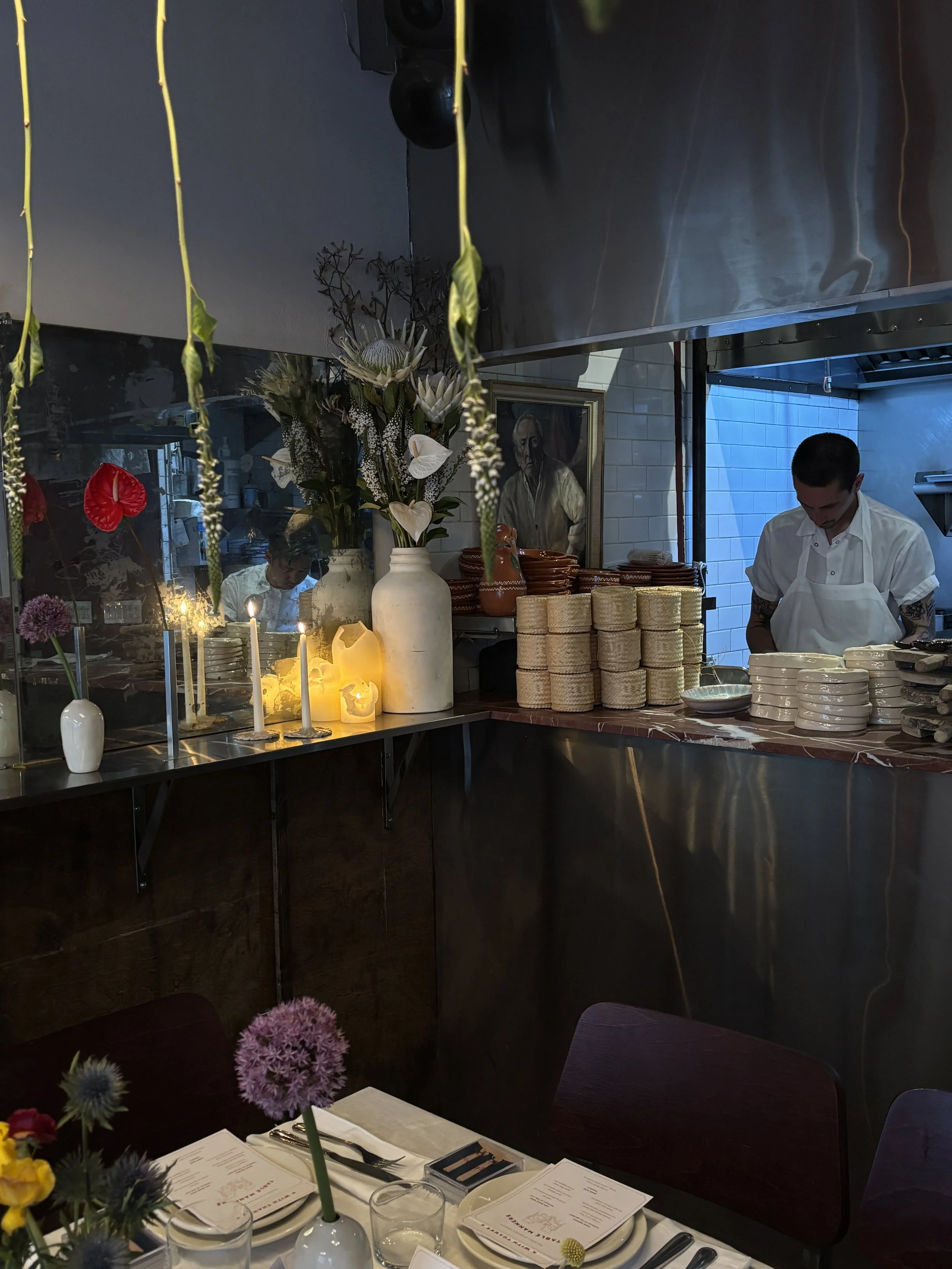 A restaurant interior with a floral arrangement, lit candles, and table settings in the foreground. In the background, kitchen staff are preparing food, with one chef visible and a portrait on the wall.