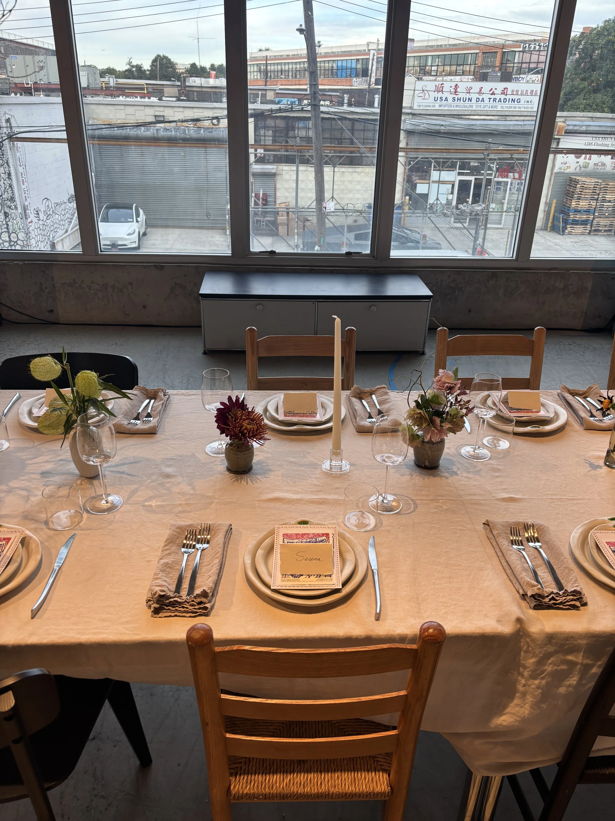 A dining table set for a meal with plates, utensils, glasses, napkins, small flower arrangements, and a tall candle in the center. The table is positioned near large windows showing an urban street with parked cars, commercial buildings, and power lines outside.