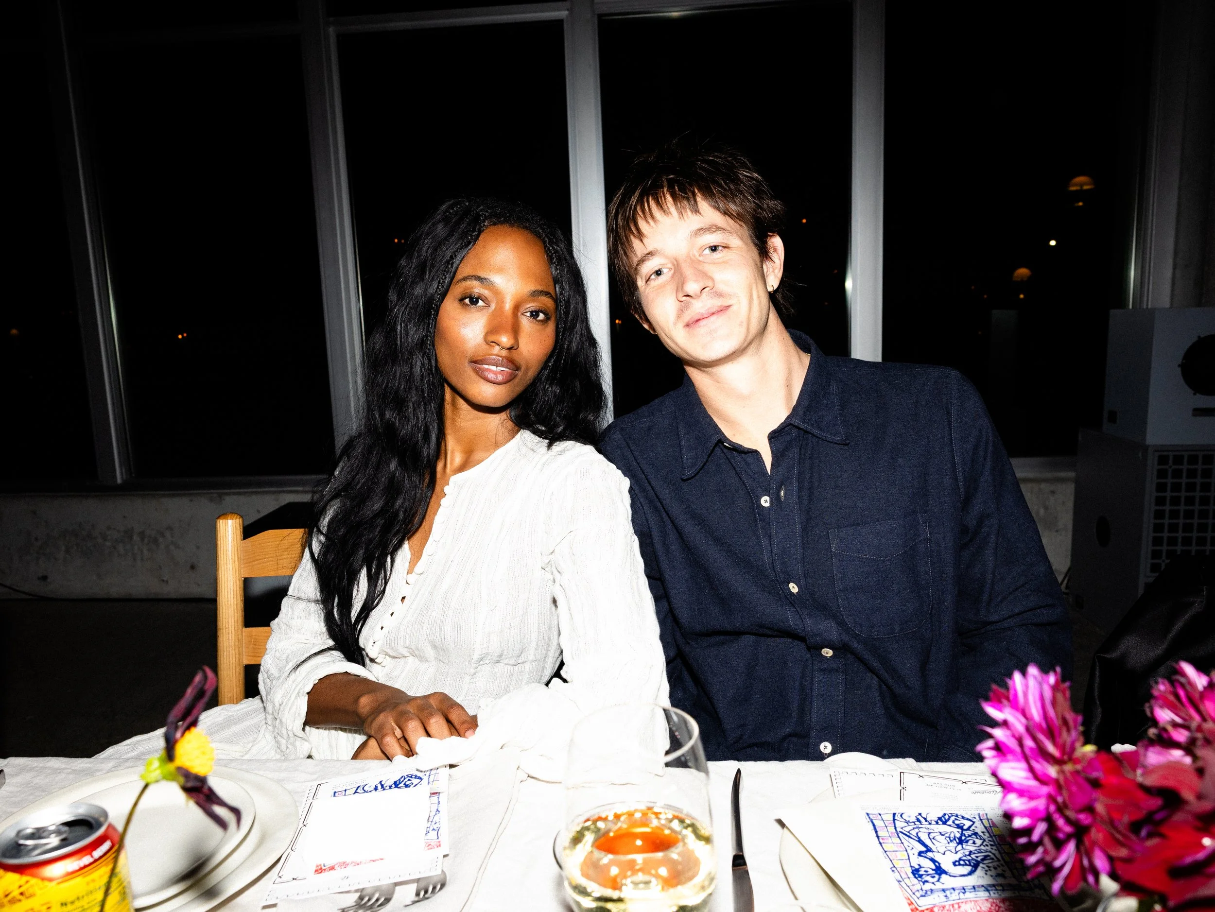 A woman and a man sitting at a table with pink flowers, drinks, and papers, with a dark night view outside the window in the background.