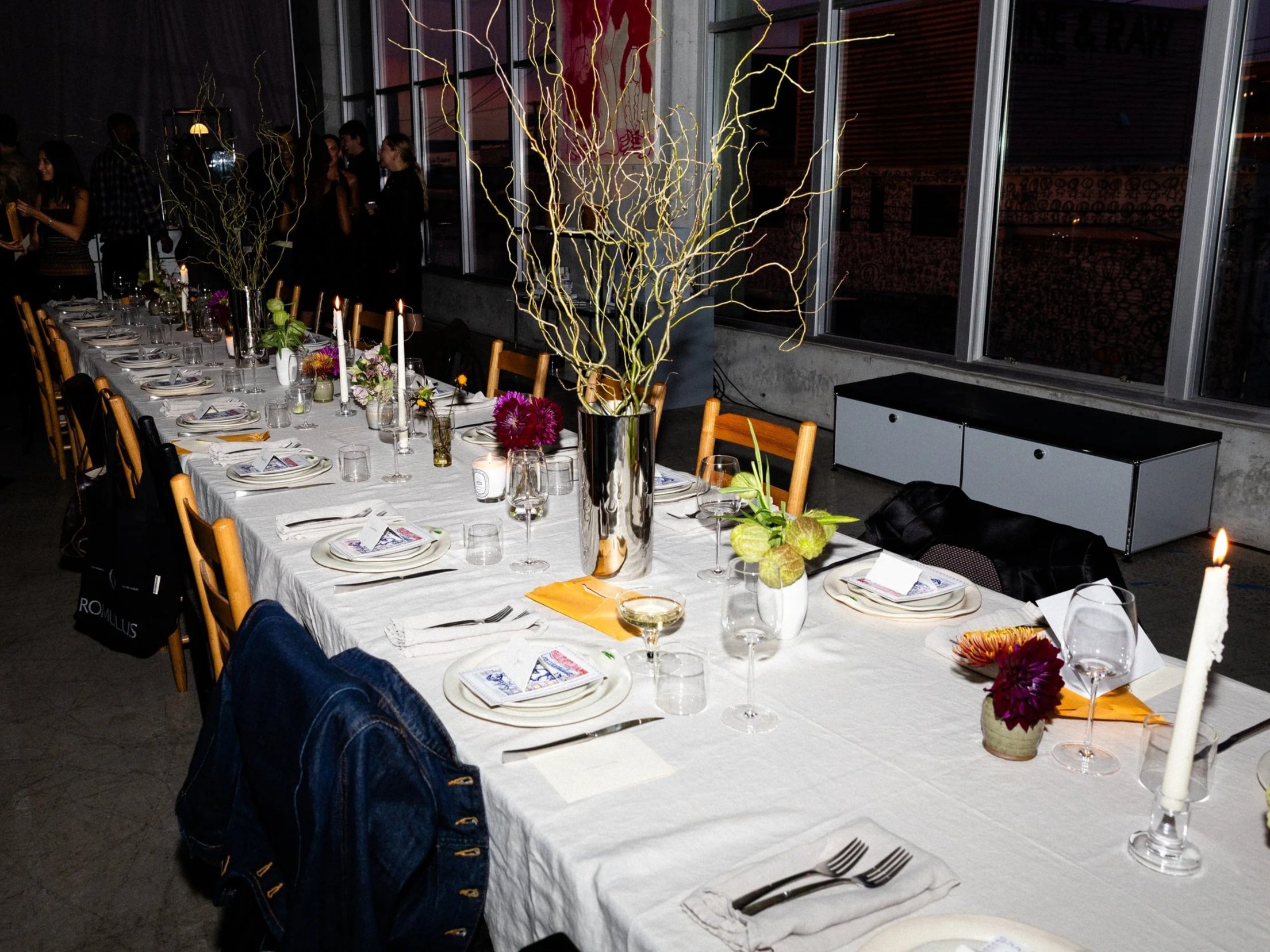 A long dinner table set with plates, glasses, cutlery, candles, and floral centerpieces, in a dimly lit modern restaurant with large windows and tall decorative branches in vases.