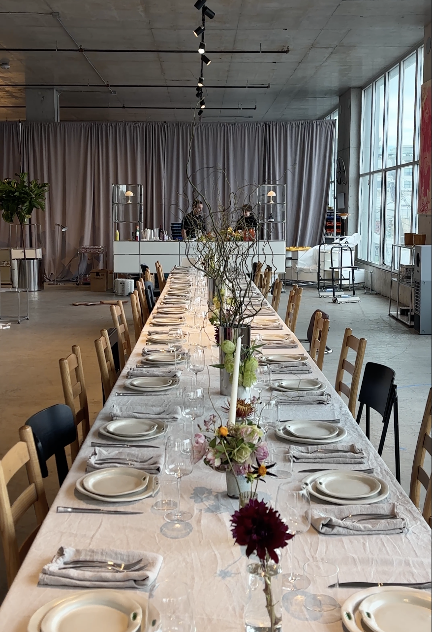 Long banquet table decorated with flowers, set with plates, glasses, and napkins in a modern, industrial-style event space with large windows and a bar area in the background.