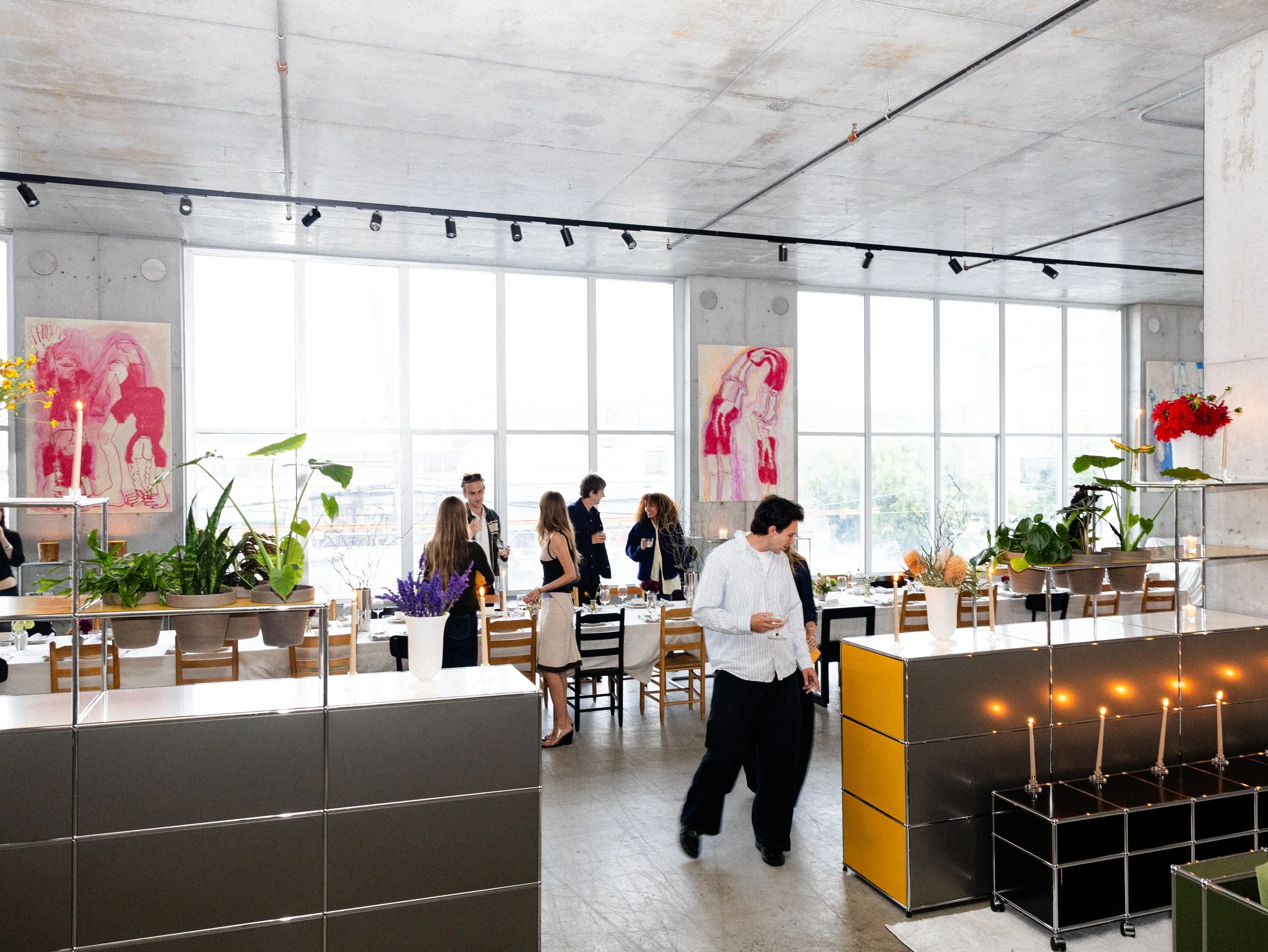 People gather in a spacious modern room with large windows, concrete ceilings, and contemporary decor, engaging in conversations around tables decorated with flowers and candles.