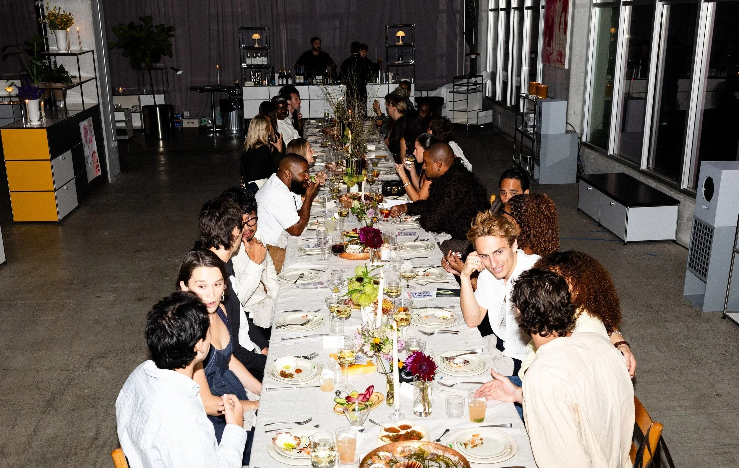 People sitting around a long dinner table at a social gathering or celebration, with drinks and floral centerpieces, in an indoor space with large windows and modern decor.