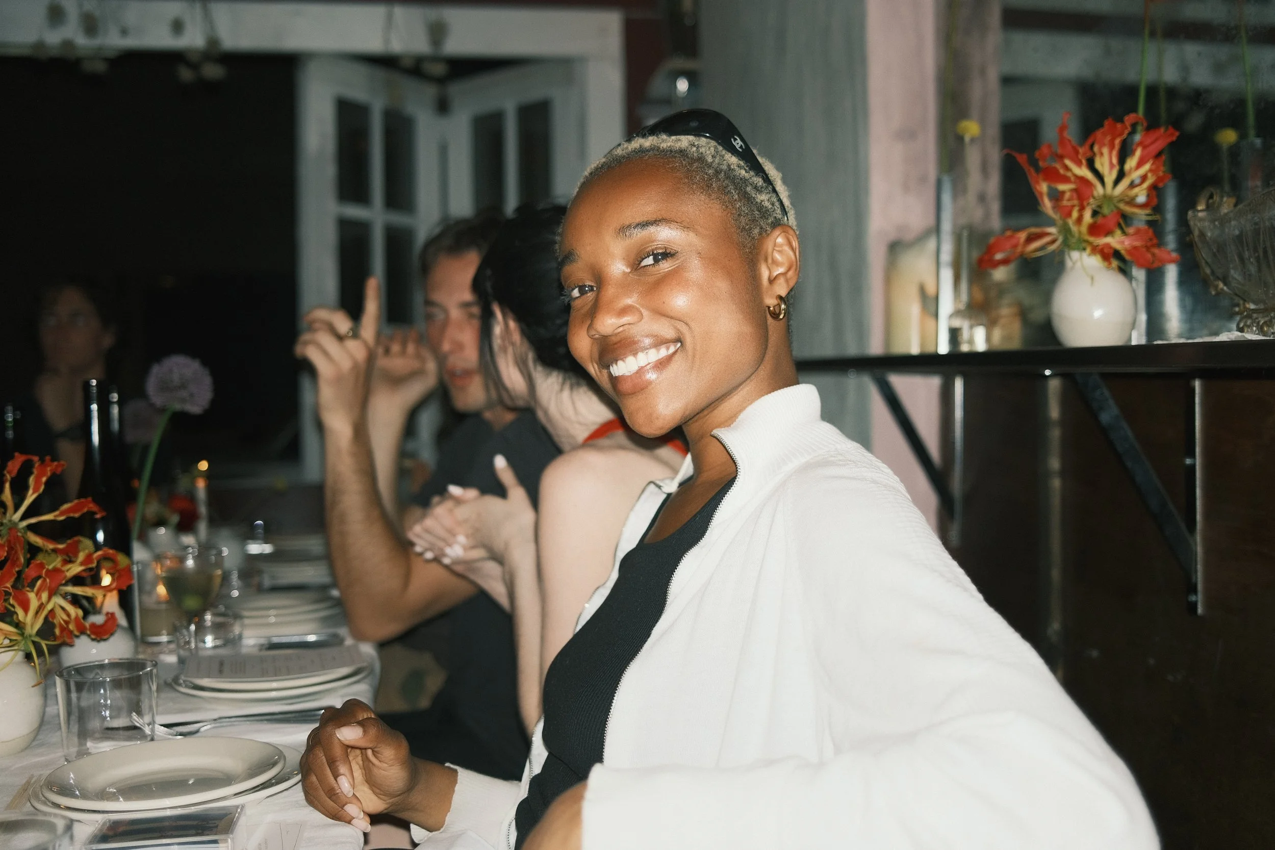 A smiling woman with short blonde hair and gold hoop earrings sitting at a long table with her friends in a cozy dining setting. The table is set with plates, glasses, and a vase of red flowers.