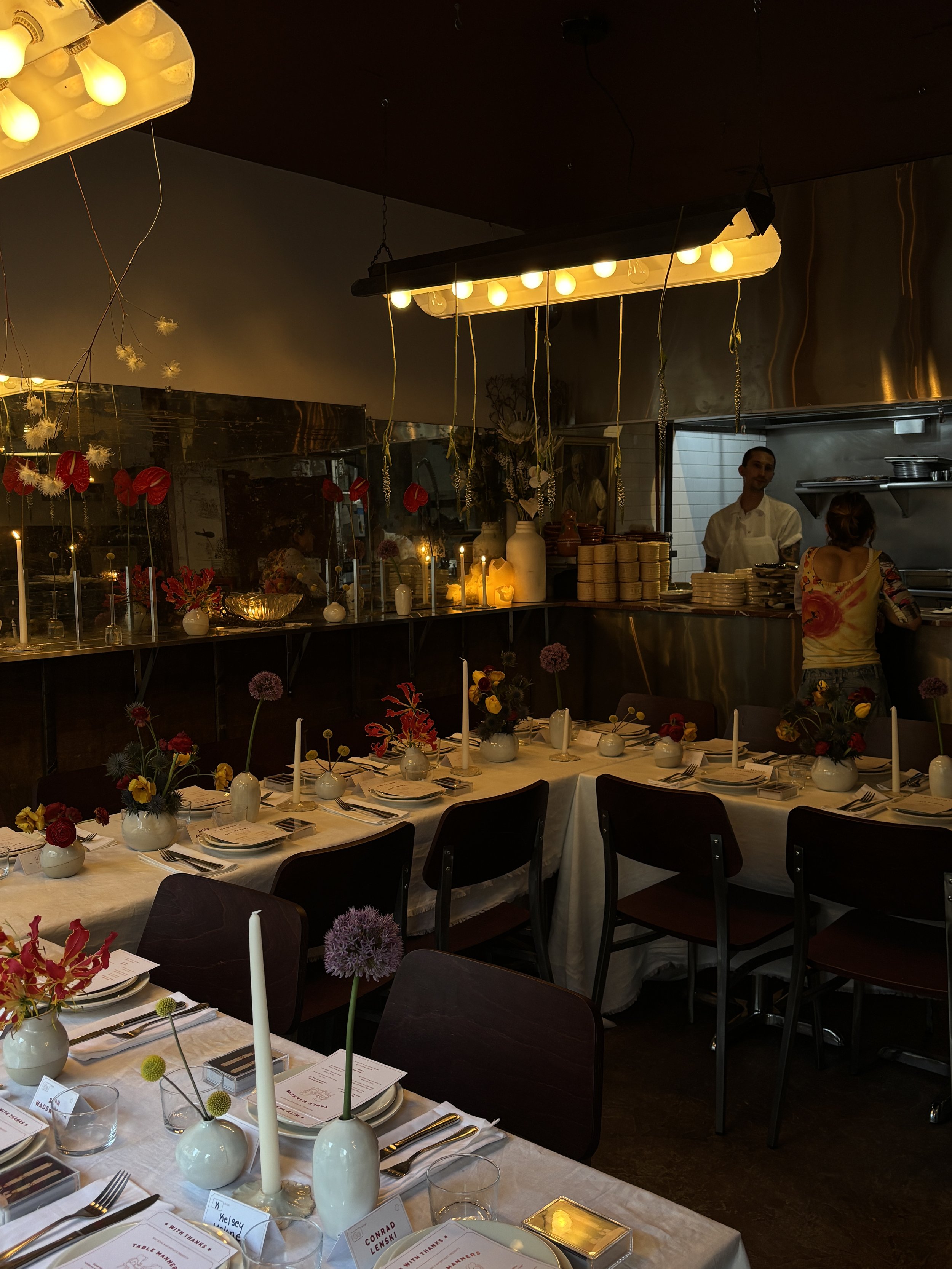 A restaurant decorated for a special occasion with tables set with white tablecloths, flower arrangements, candles, and place cards. There are hanging light fixtures and decorations, with a kitchen area and staff in the background.