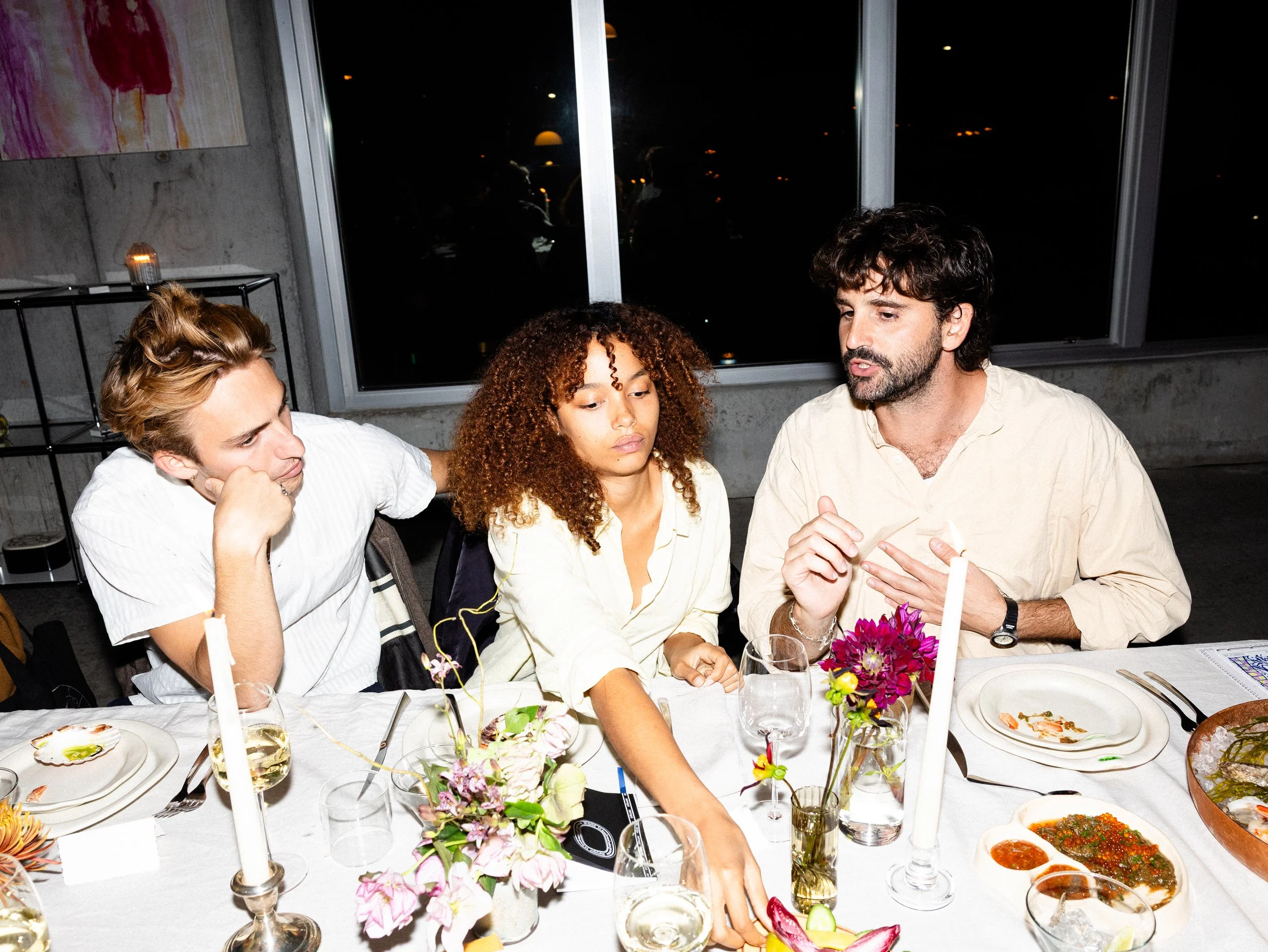 People sitting at a dinner table with floral centerpieces, candles, and plates of food, engaged in conversation at a nighttime gathering.