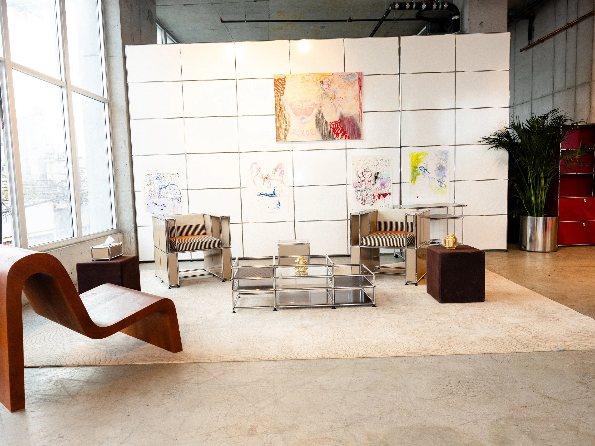A modern interior with a white tiled wall displaying colorful abstract artwork, chairs with bar seating, a large window on the left, and a potted plant on the right.