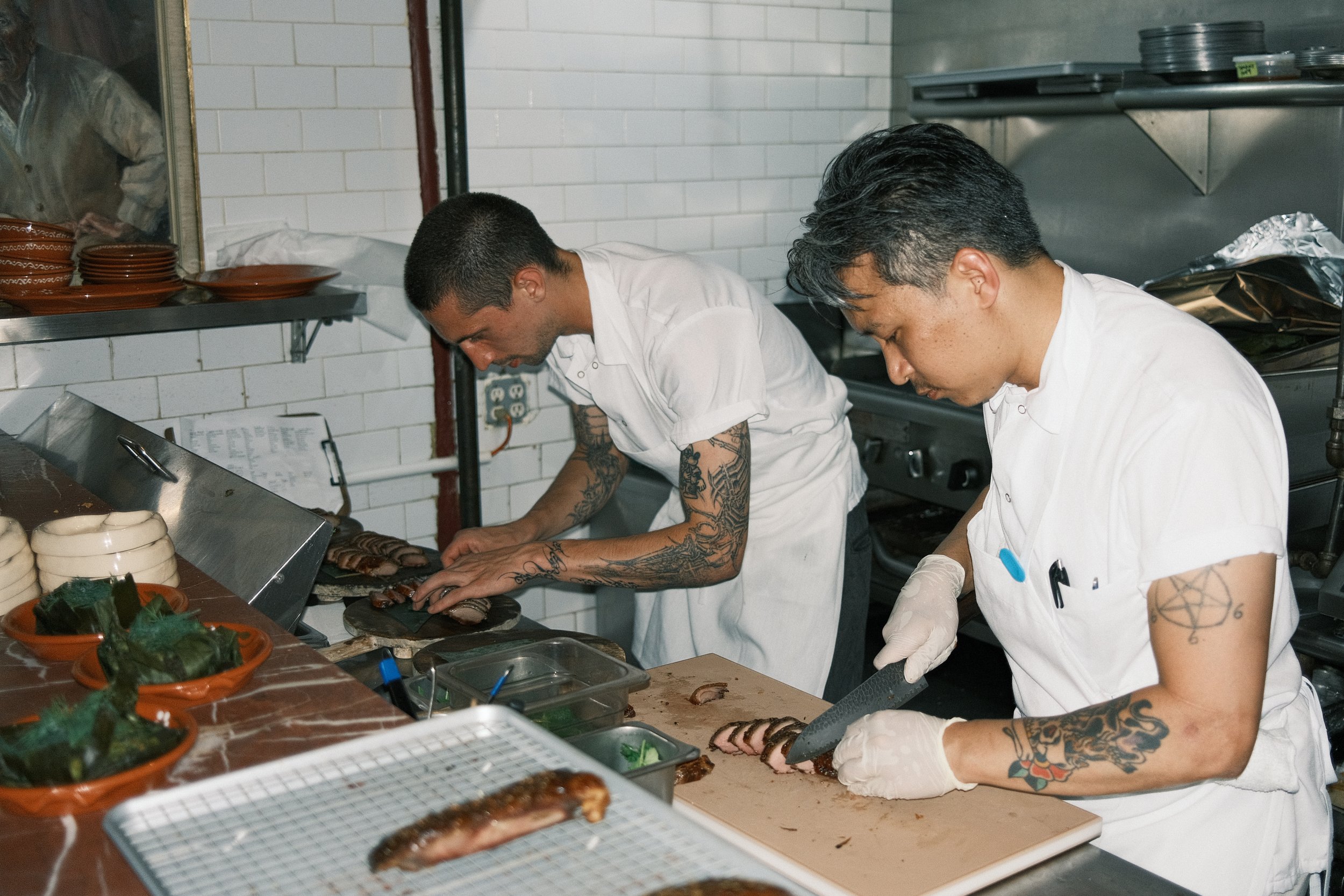 Two chefs, both with tattoos, are preparing meat in a professional kitchen. They are focused on slicing the cooked meat on a cutting board, wearing white chef coats and gloves. The kitchen has white tiled walls, and there are plates, bowls, and kitchen tools visible around them.