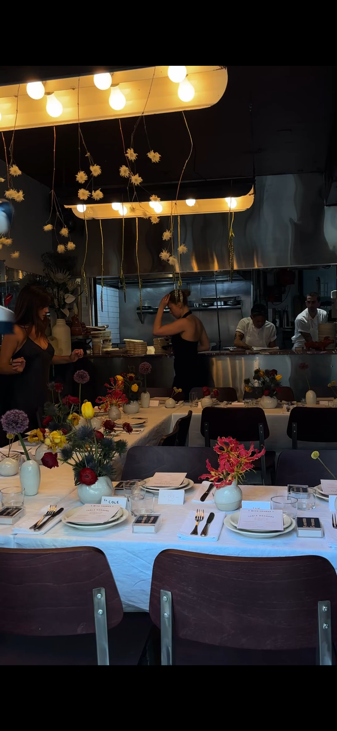 A restaurant interior with a long table set for dining, decorated with vases of colorful flowers, and a view of the open kitchen with staff preparing food under hanging lights and decorative flowers.